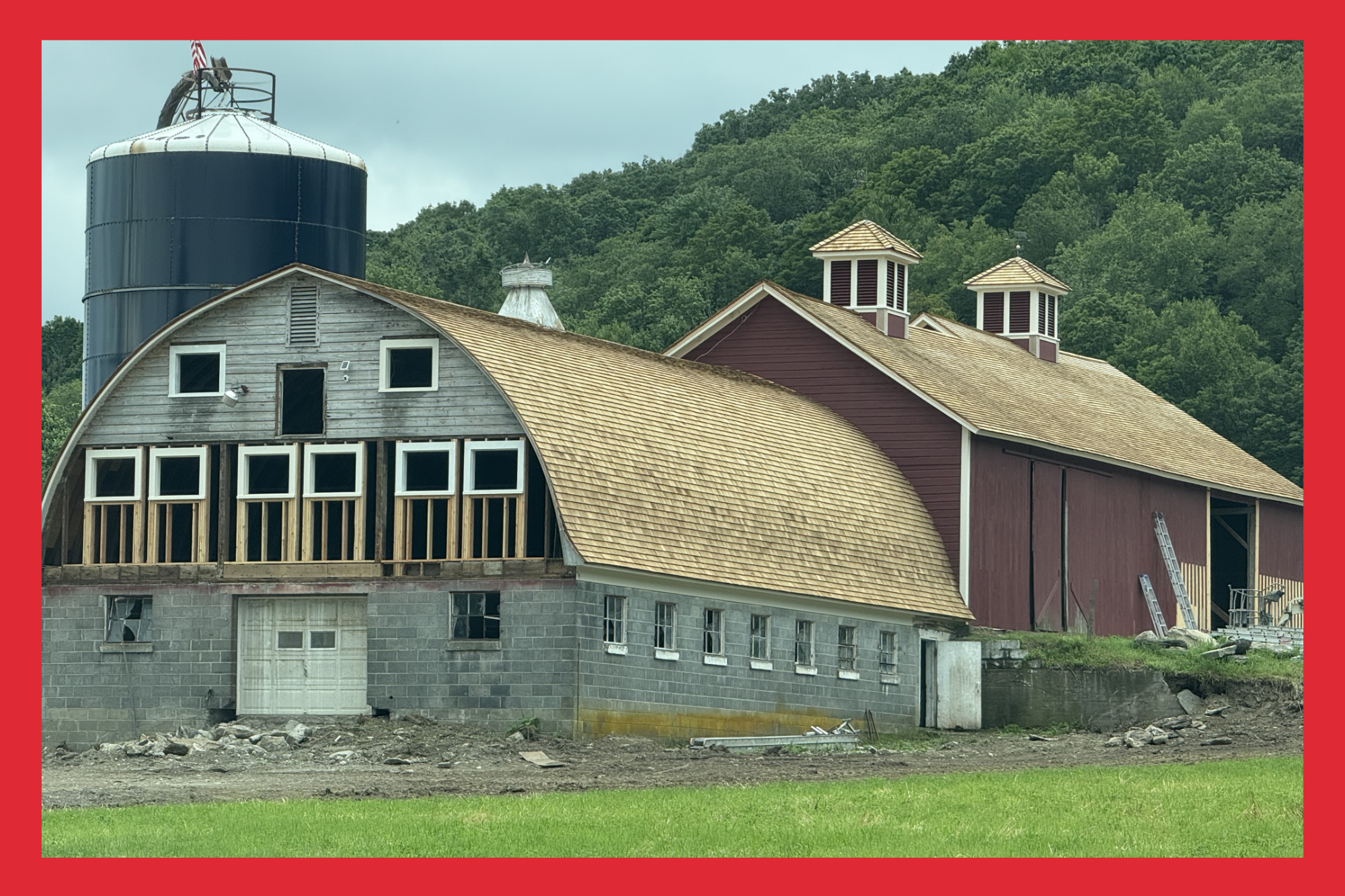 Wrapping construction site of a barn, with a new curved roof, windows, a large door, and a silo in the background, surrounded by green hills.