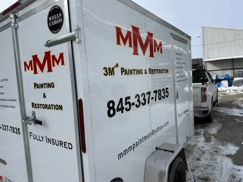 White mobile trailer with branding for MMM Painting & Restoration, including company name, contact number, and email address, parked outside in a snowy area.