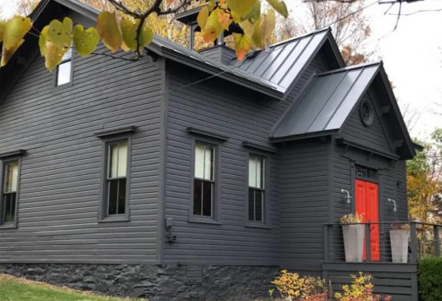 A modern, dark gray house with a bright red front door, multiple windows, a metal roof, and a small porch with two large planters.