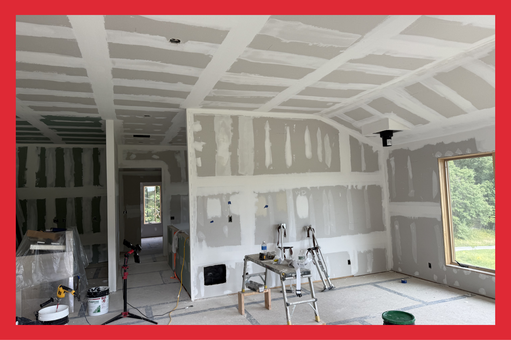 Interior of a room under construction with drywall walls, ceiling, and window, with tools and supplies visible.
