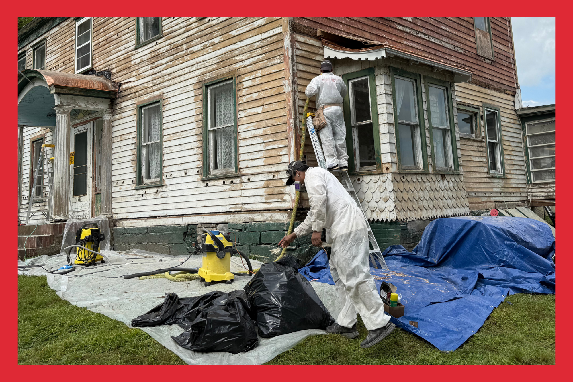 Two workers in white protective suits restoring the exterior of an old, weathered wooden house, with one on a ladder working on the upper wall and the other on the ground handling tools and materials, surrounded by cleaning equipment and tarps.