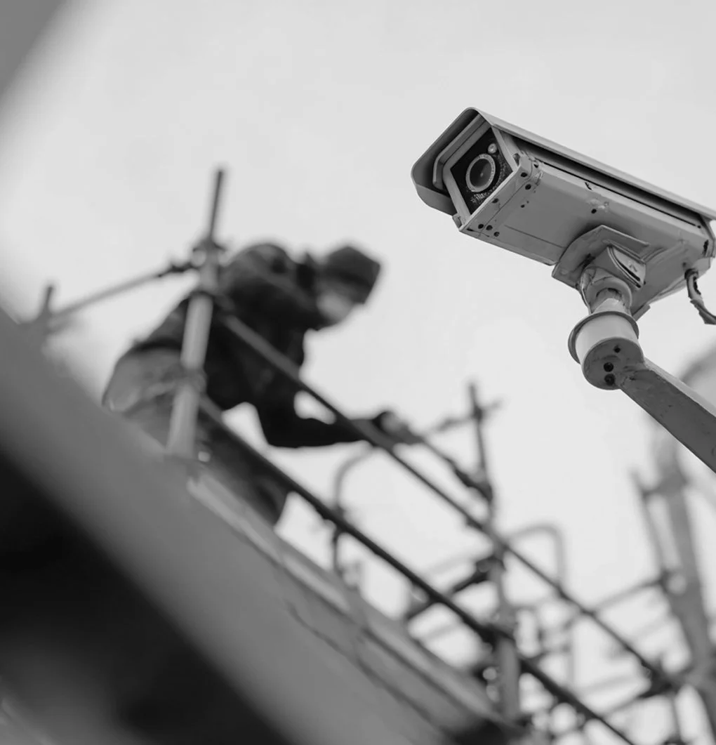 Close-up of a security camera mounted on a pole with a construction worker working on scaffolding in the background.