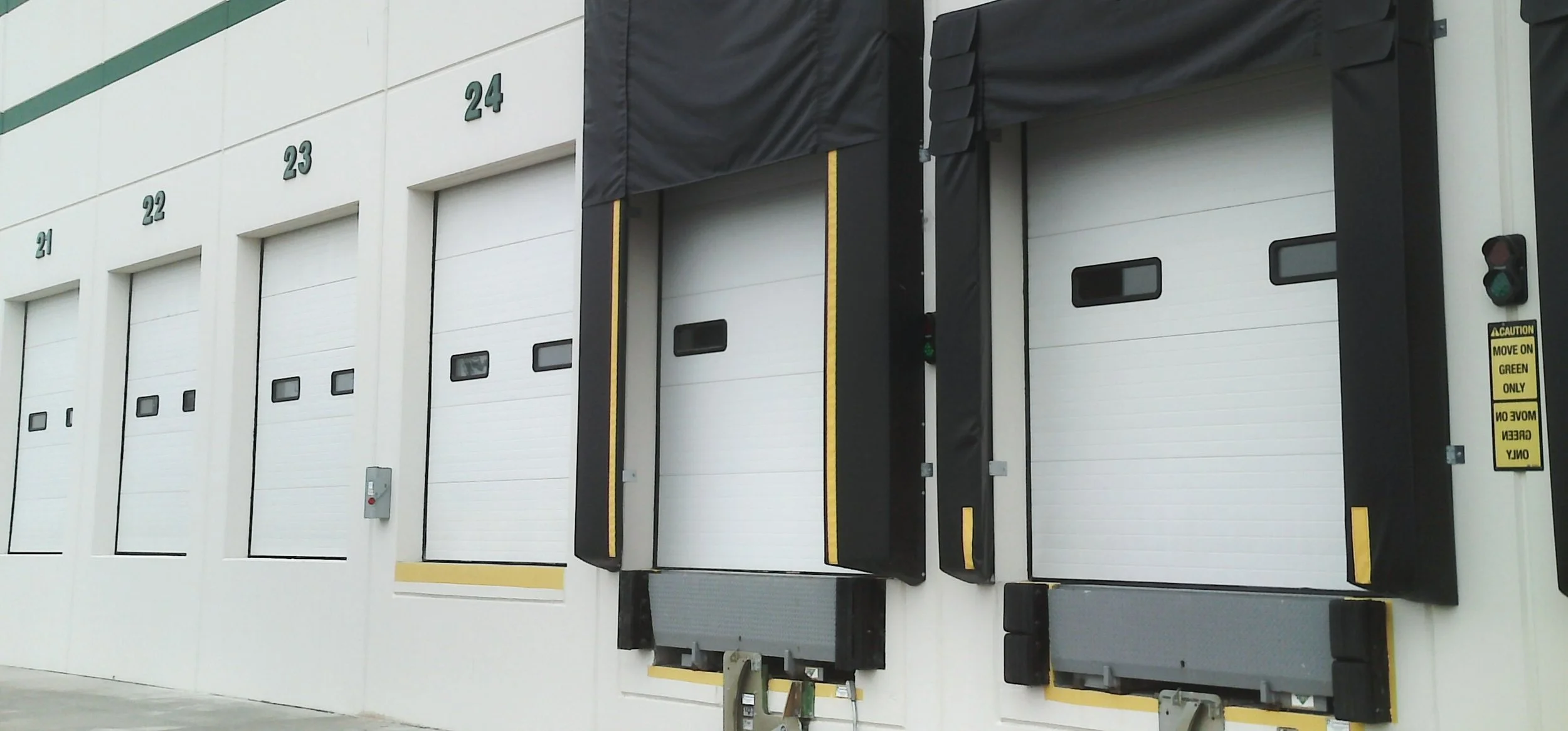 Loading dock with multiple white roll-up doors numbered 21 to 24, each with black trim and small windows. Two dock levelers are visible, with caution signs and green lights beside each door.
