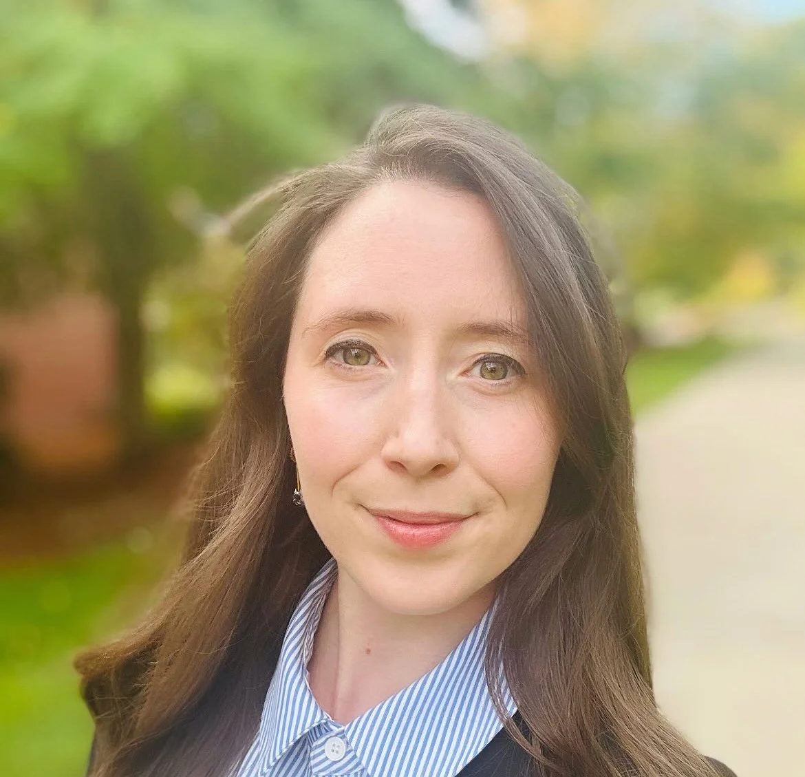 A woman with long brown hair and light skin smiling outdoors, wearing a blue and white striped shirt, with a blurred green and yellow background.