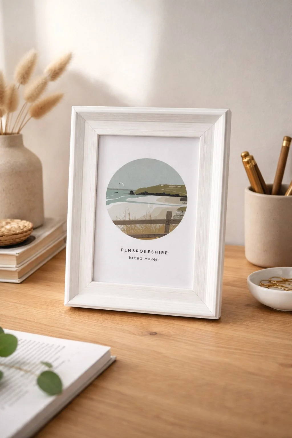 Framed art prints of Broad Haven coast in Pembrokeshire on a desk with decorative items nearby.