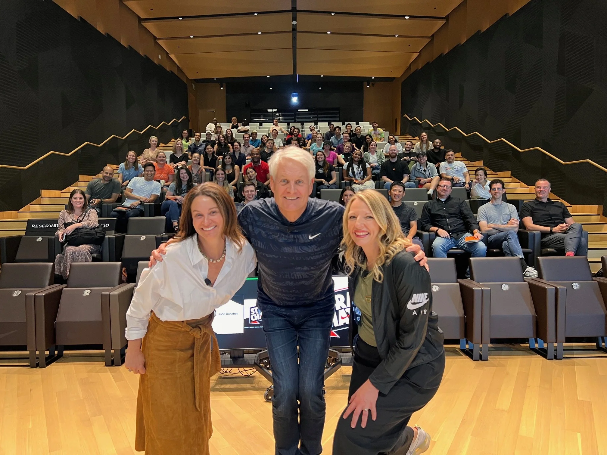 Group photo of three people in the foreground with a large audience seated behind them in an auditorium. The setting appears to be a presentation or event, with all subjects smiling and posing for the camera.
