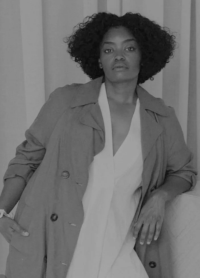 A woman with curly hair wearing a trench coat over a light dress, standing indoors against a wall with vertical paneling, looking confidently at the camera.