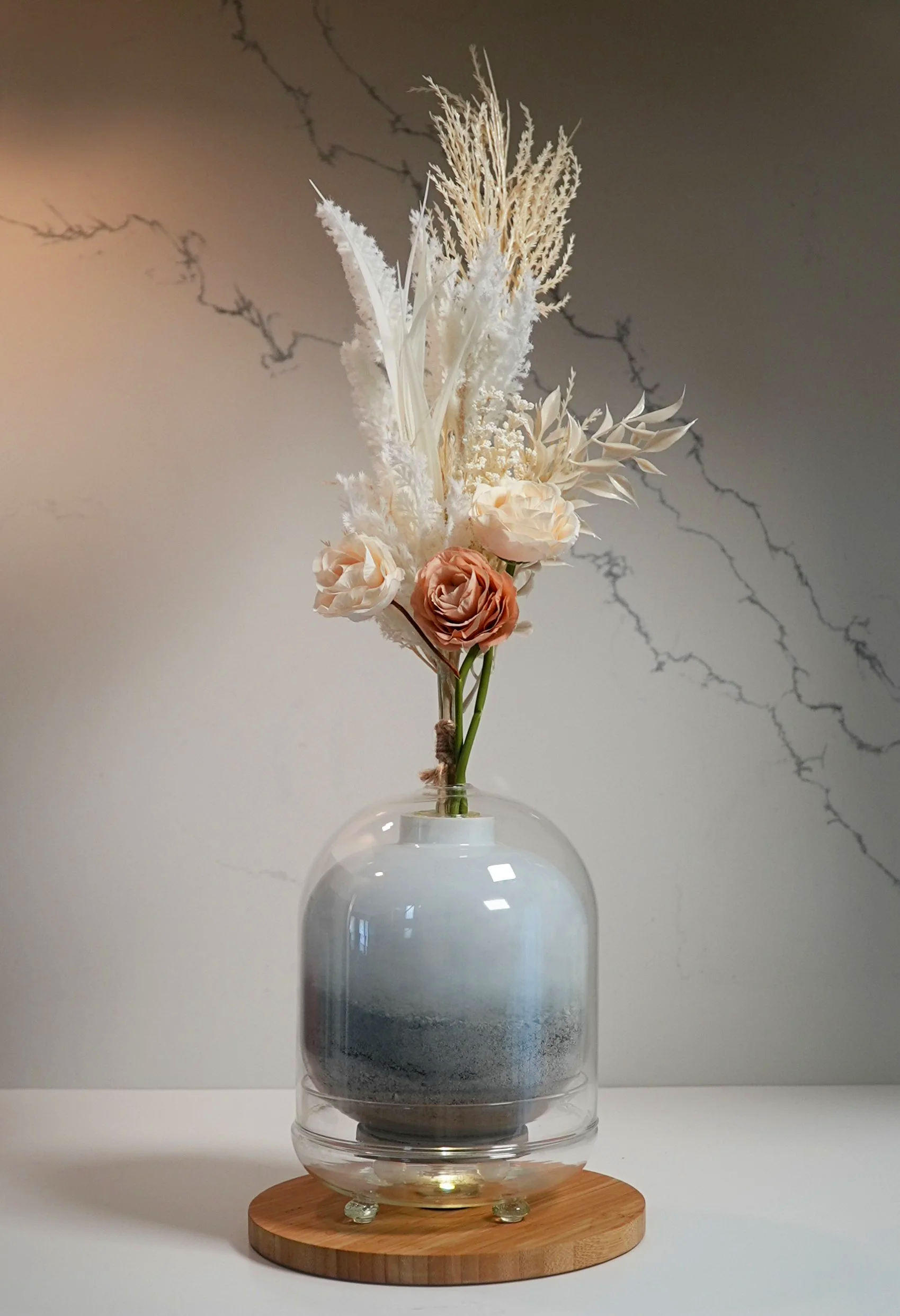 Custom Urn - Decorative bouquet of dried flowers in neutral tones, arranged in a clear glass vase with a rounded shape, placed on a wooden base against a light-colored wall.