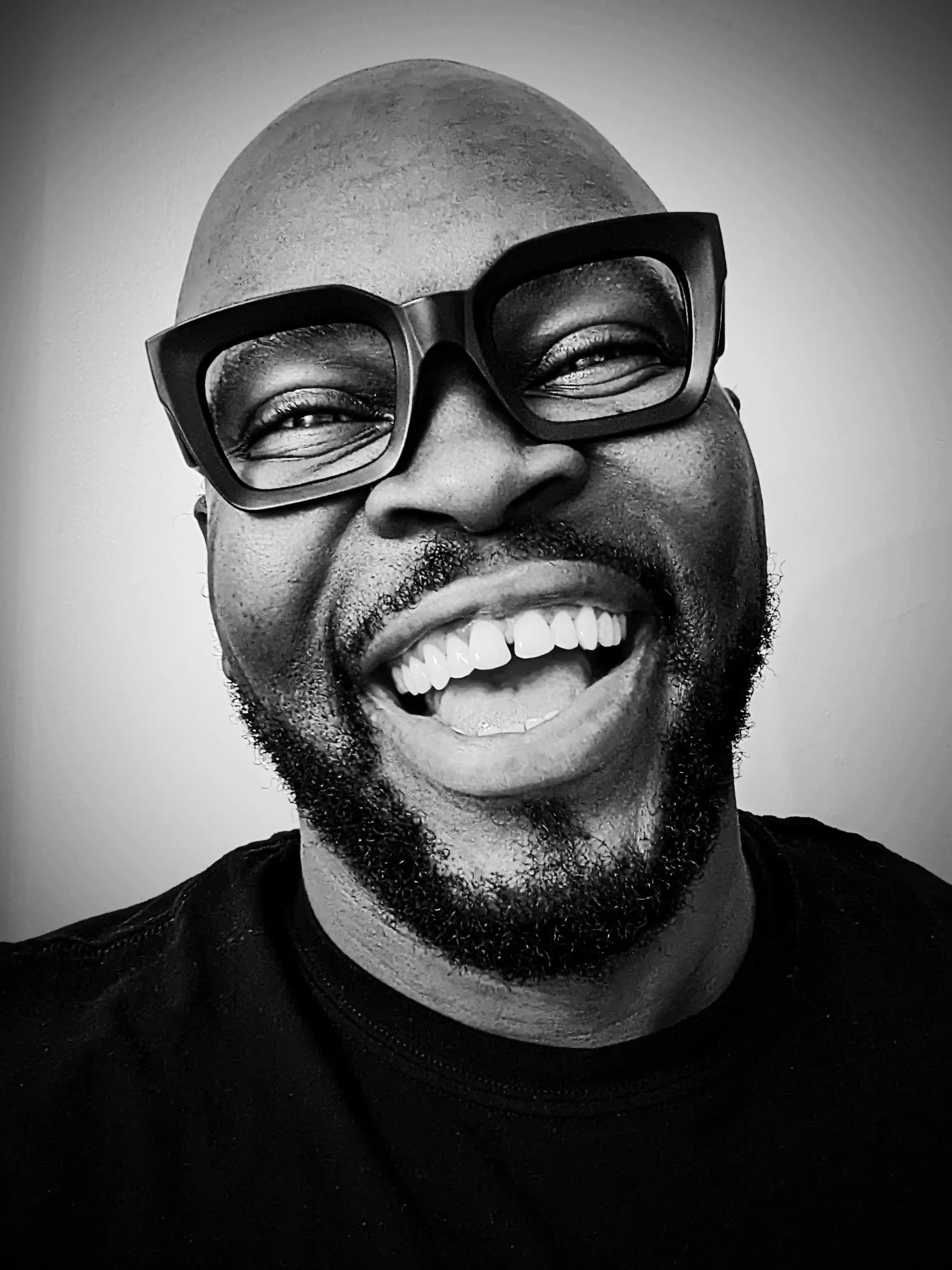 A black and white close-up photo of a man with a bald head, wearing large glasses, smiling widely showing his teeth, with a beard and mustache, against a plain background.