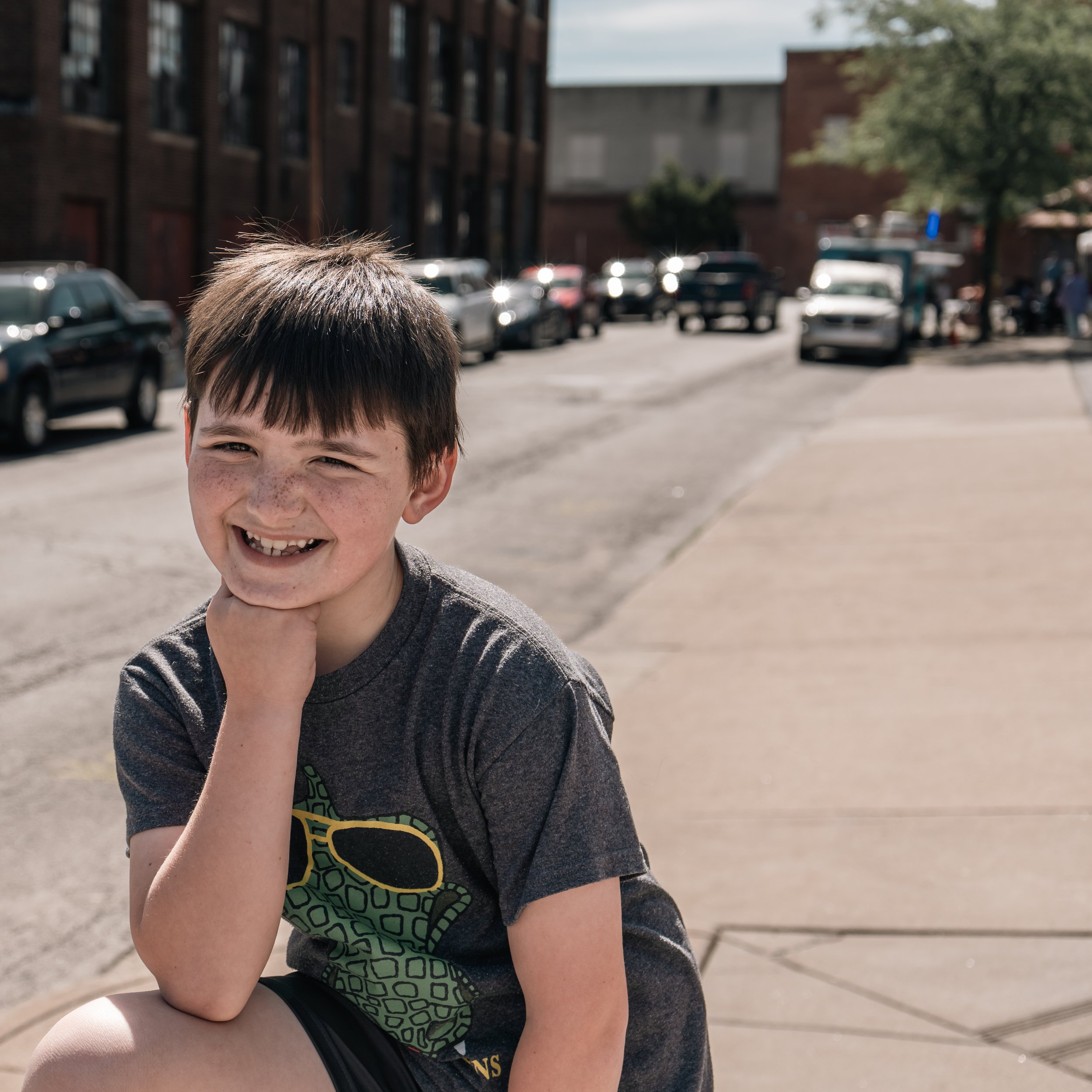 A young boy with short brown hair and freckles, smiling and resting his chin on his hand, sitting on a sidewalk in an urban area with parked cars and buildings in the background.