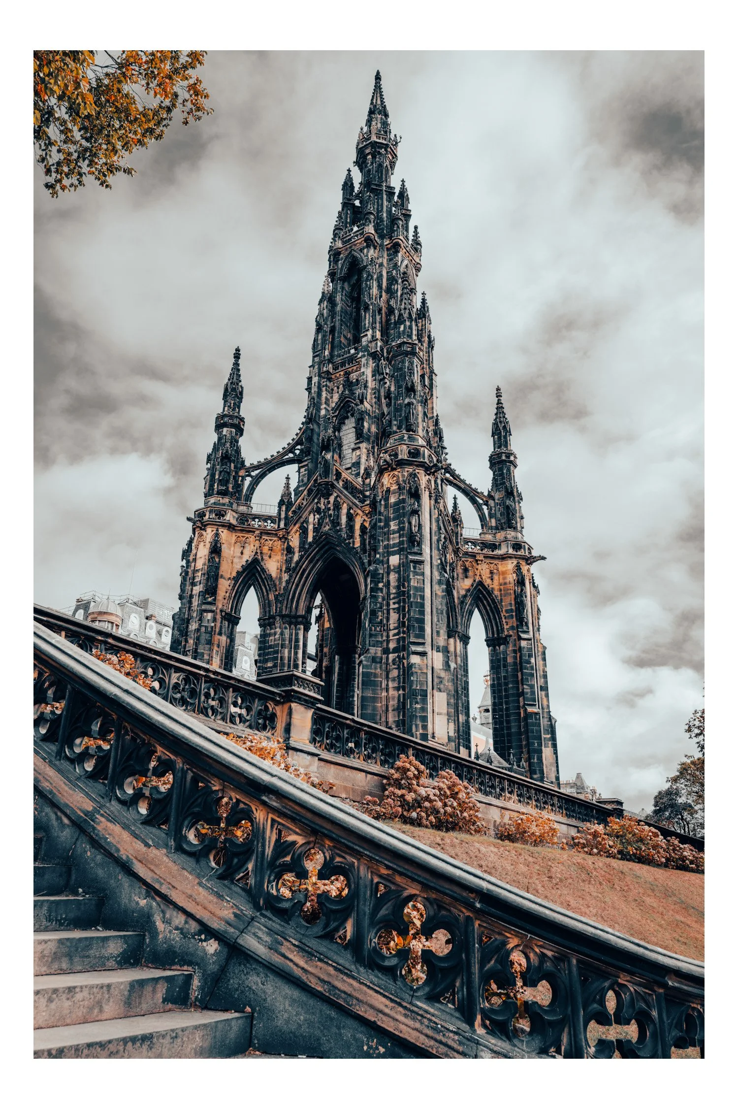 Gothic-style dark stone church with tall spires, viewed from the stairs with decorative iron railing, under cloudy sky with some autumn leaves on the ground.