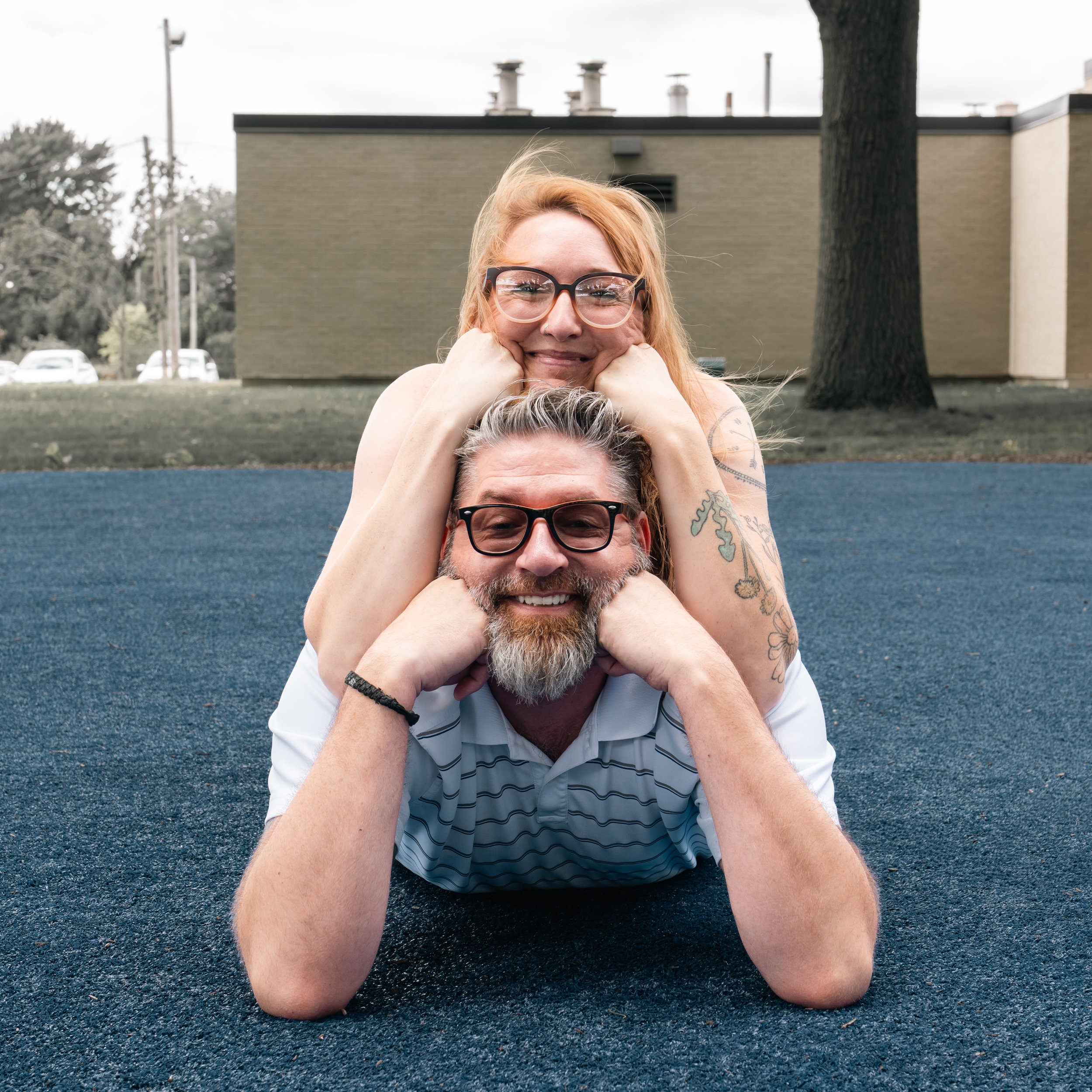 A smiling couple, a woman with glasses and tattoos, on top of a man with glasses and a beard, lying on a blue surface outdoors.