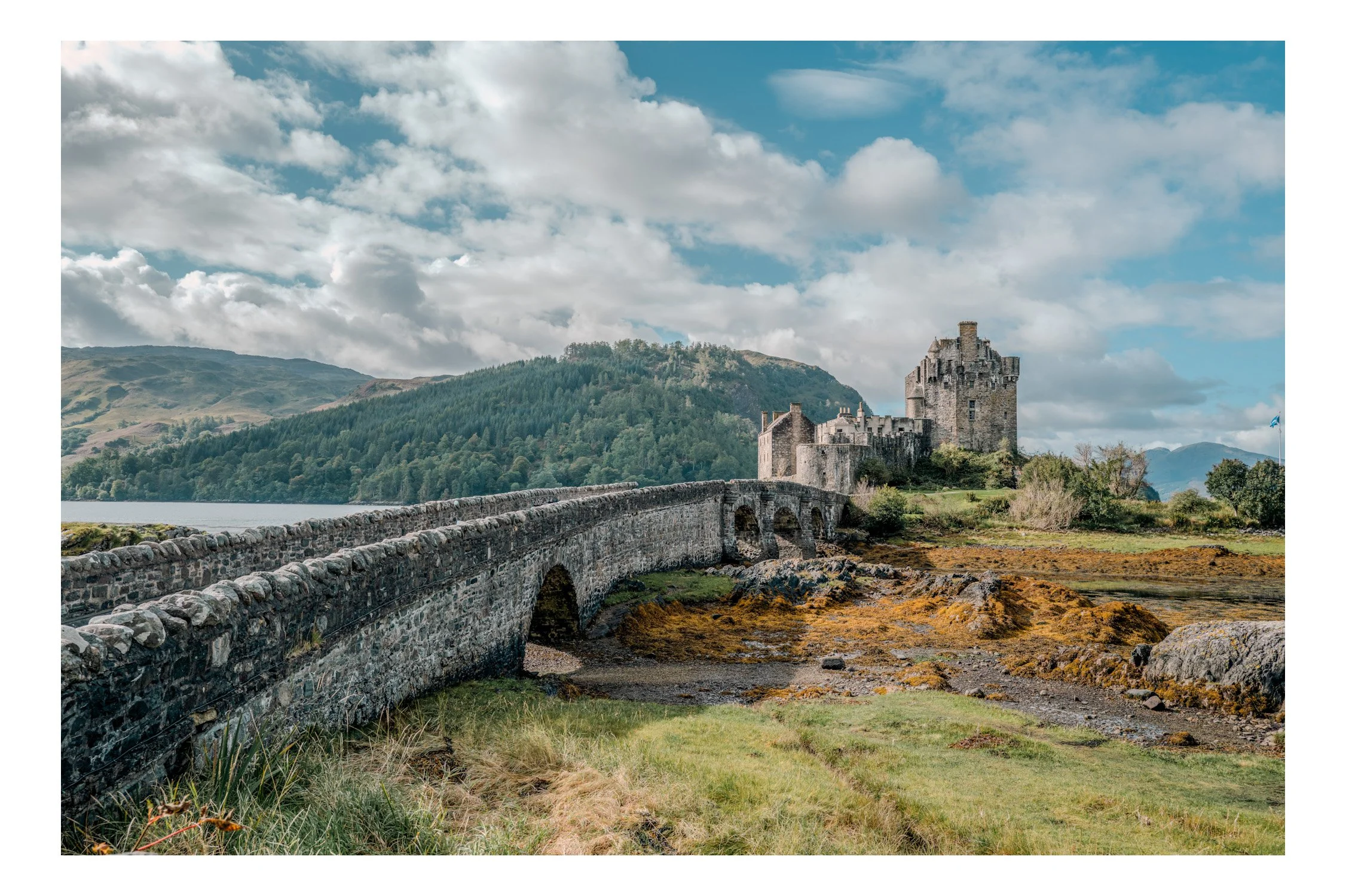 A stone bridge leading to a medieval castle on a grassy landscape with hills and mountains in the background under a partly cloudy sky.