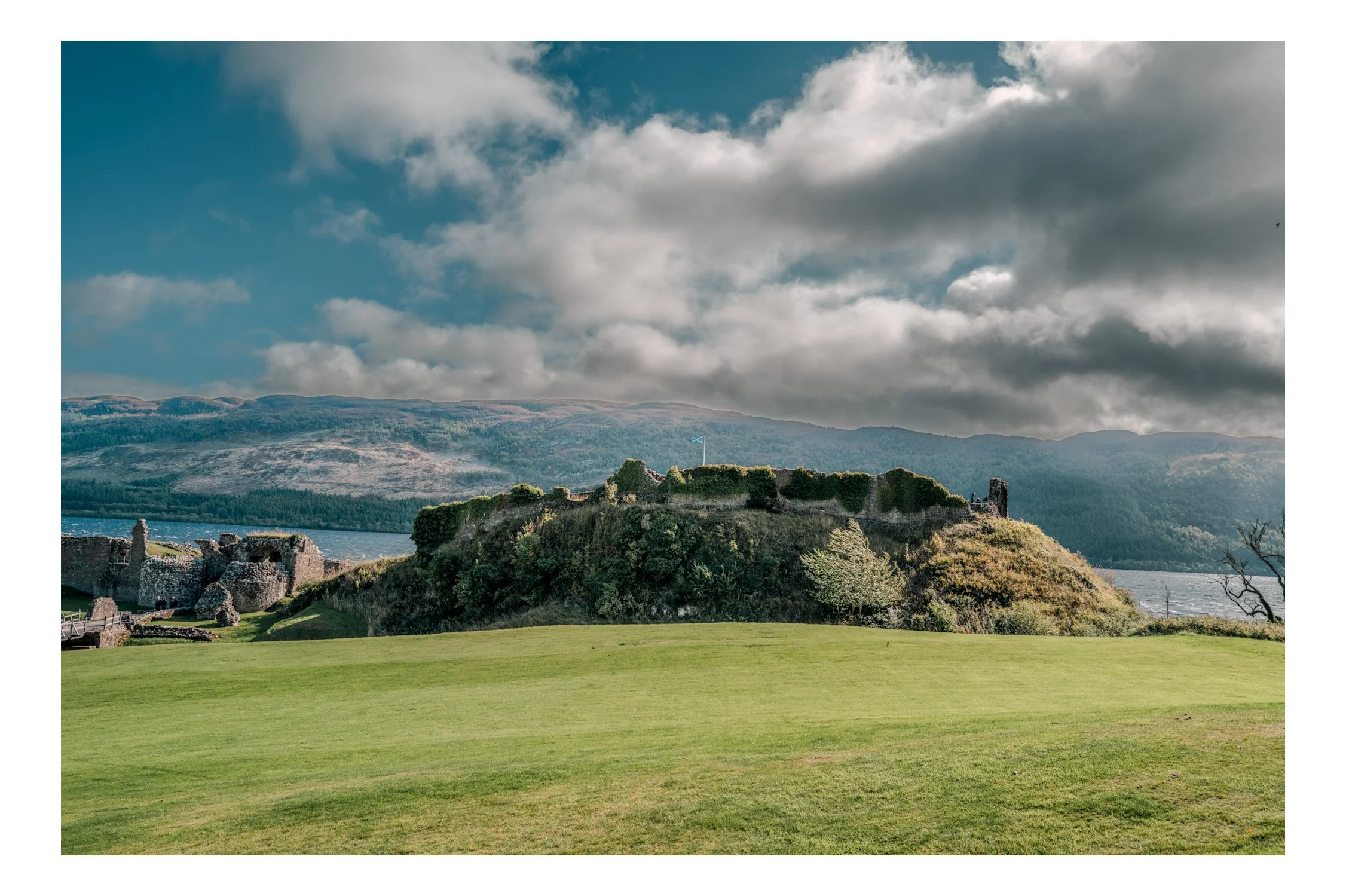 Ruins of an ancient castle on a hill with a flag, overlooking a green field, water, and distant hills under a cloudy sky.