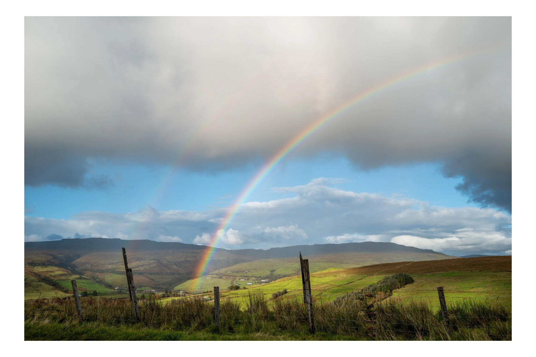 Landscape with green fields, rolling hills, and a sky filled with clouds and a rainbow.