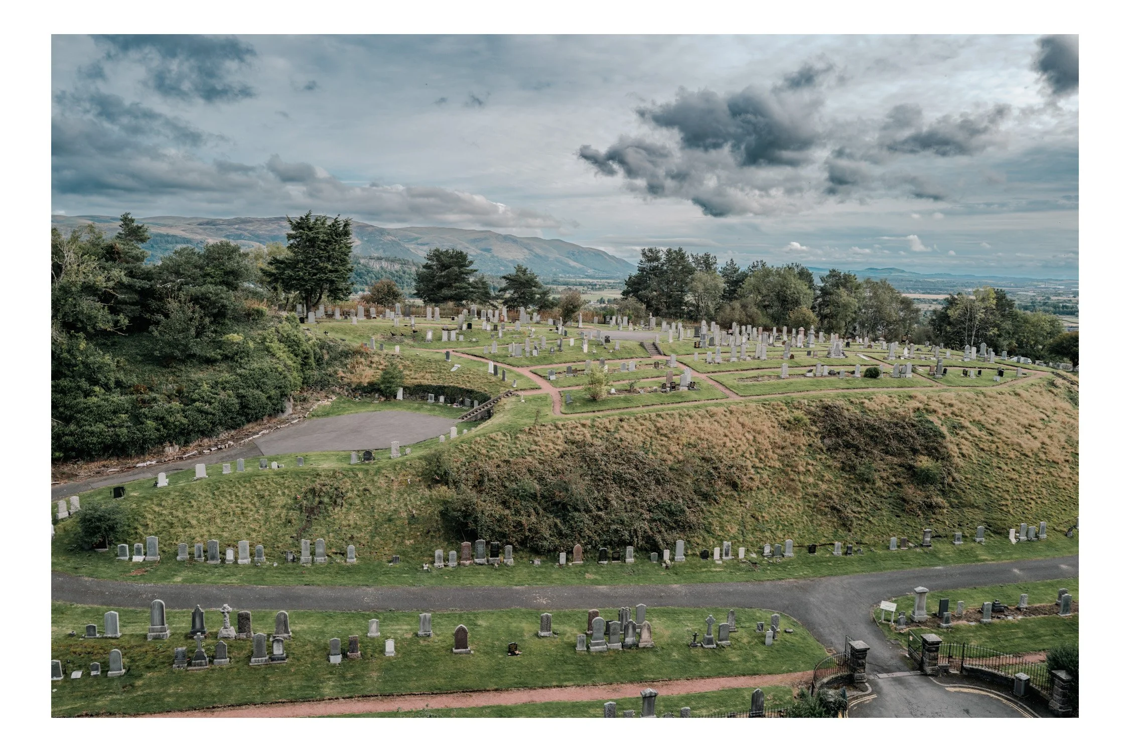A graveyard on a hill with numerous tombstones, some with crosses, surrounded by green grass and trees. A footpath winds through the graves, leading to a gate at the bottom right corner. In the background, there are distant mountains and a cloudy sky