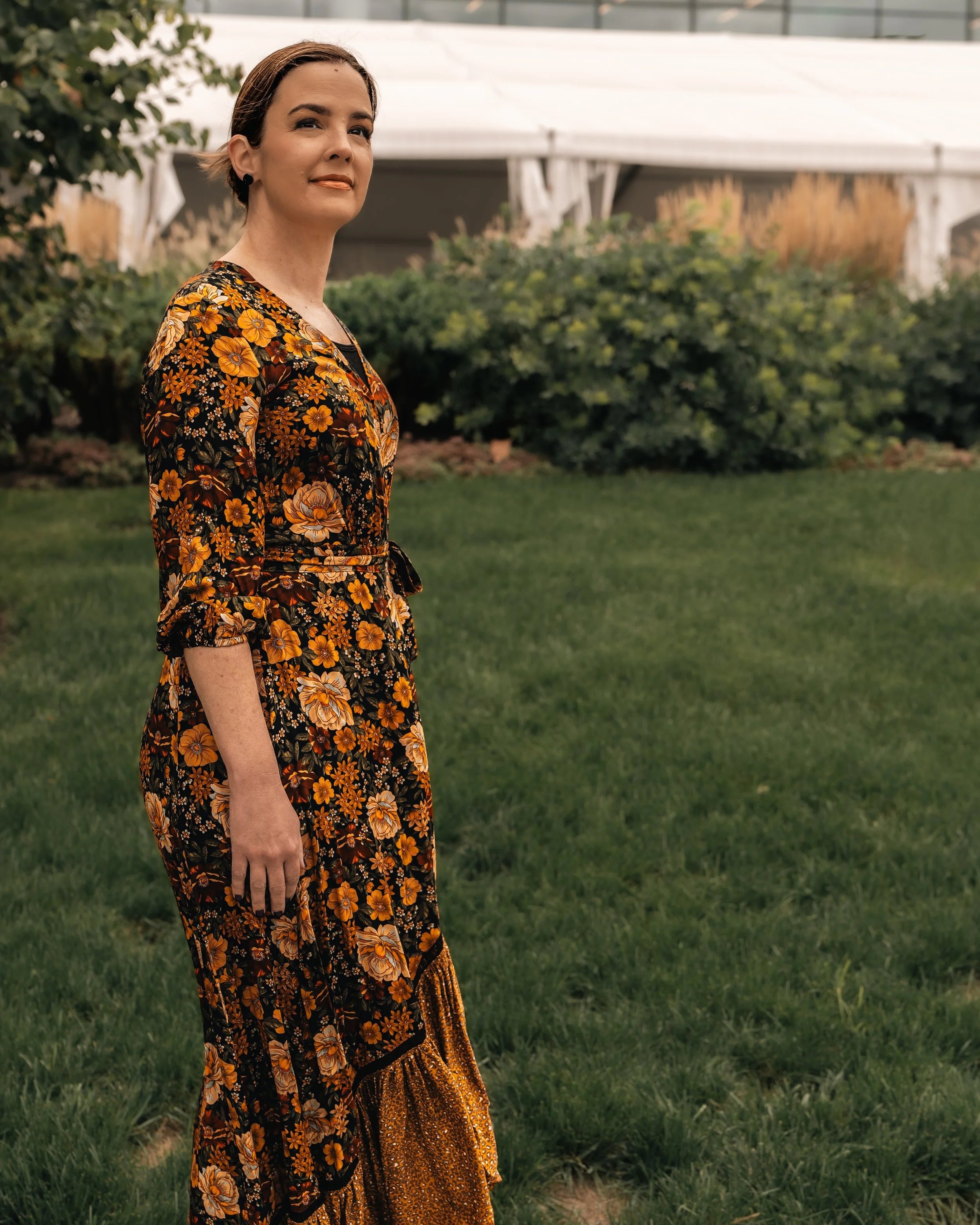 A woman in a black and orange floral dress standing outdoors on a grassy area with greenery and a white tent in the background.