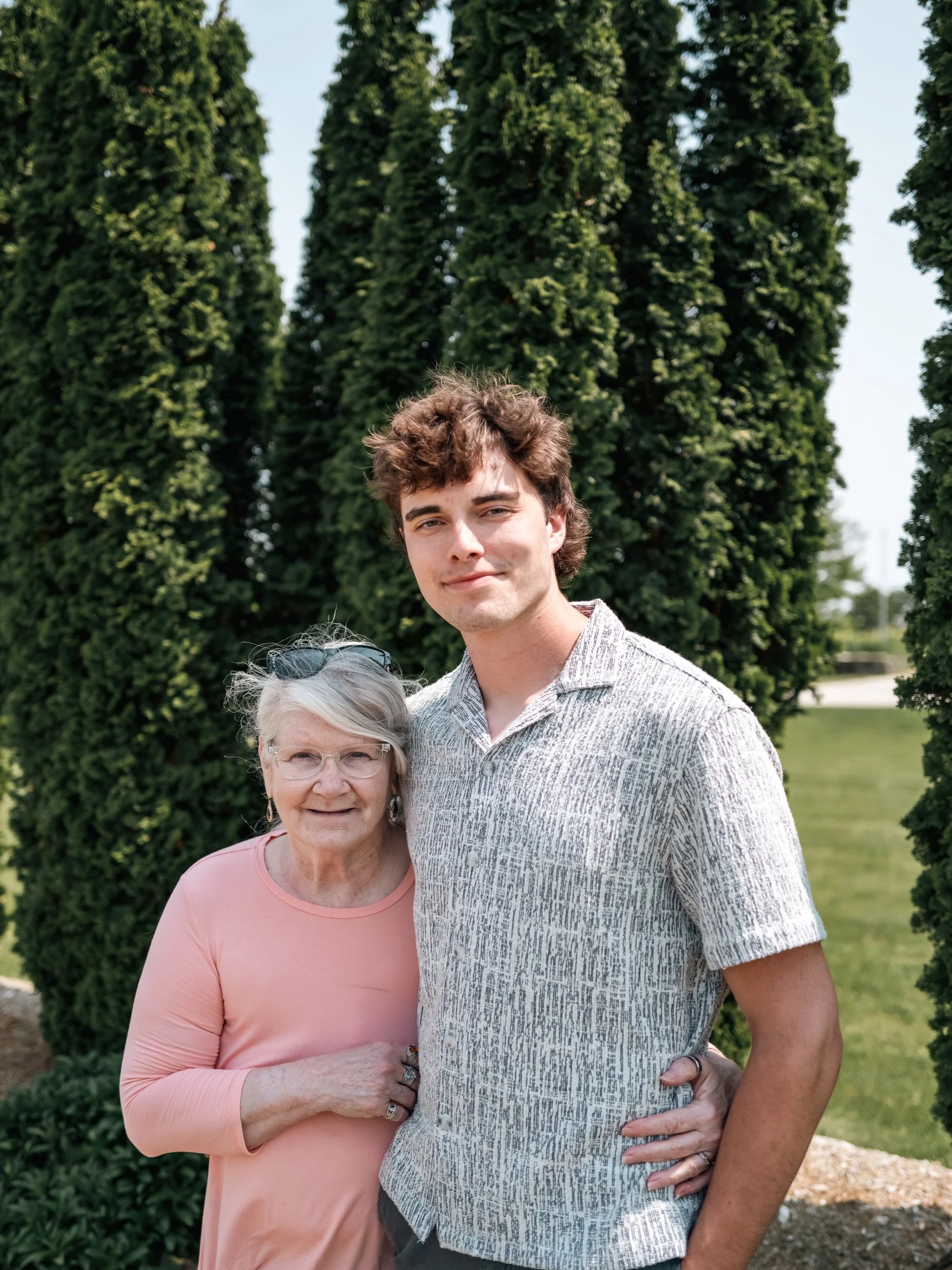 A young man and an elderly woman smiling outdoors with tall green trees in the background.