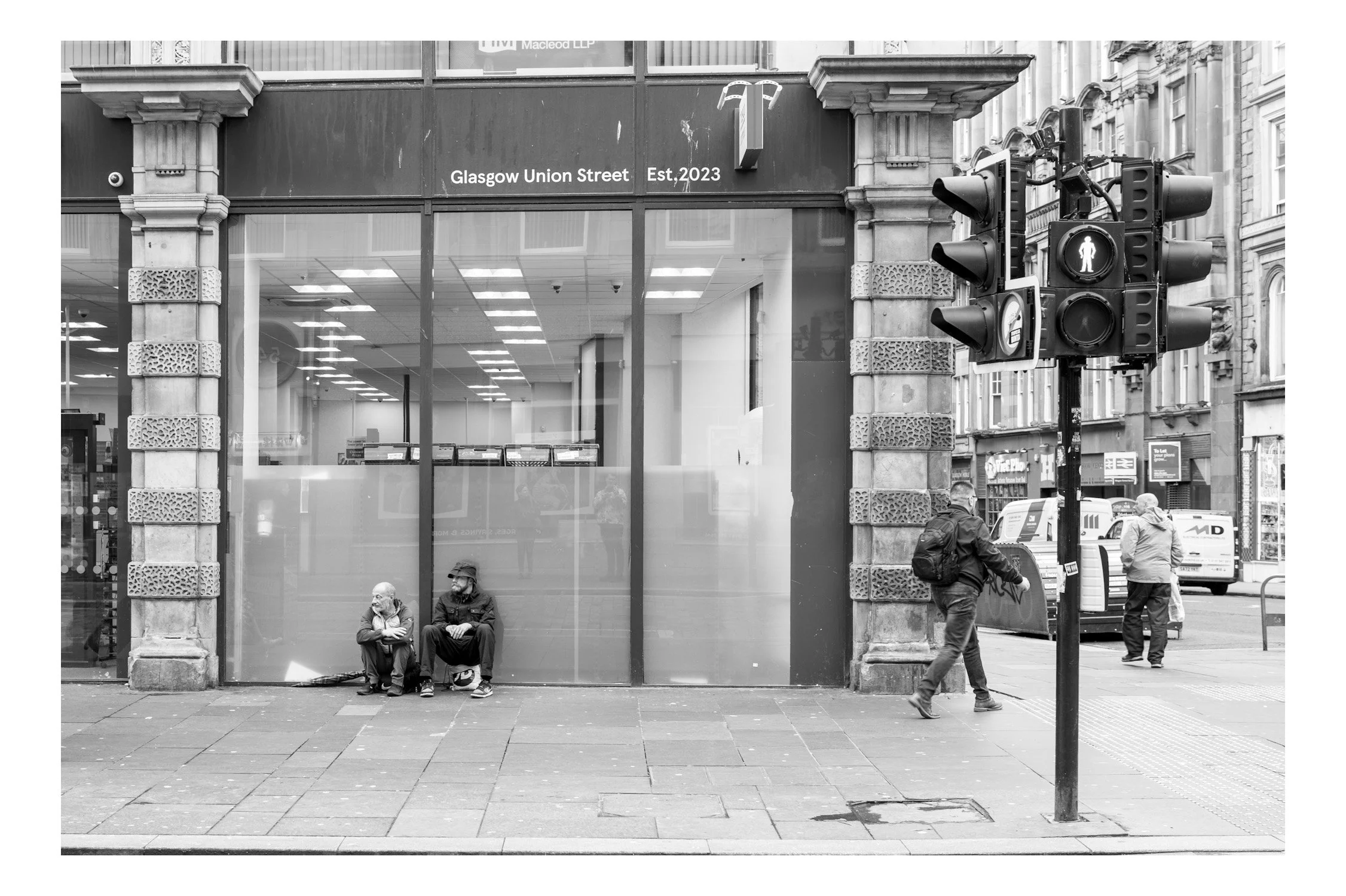 Street scene showing pedestrians waiting at a crosswalk with traffic lights in front of a building with large windows and a sign reading "Glasgow Union Street" and "Est. 2023". Two people are sitting on the sidewalk, while two others are walking near