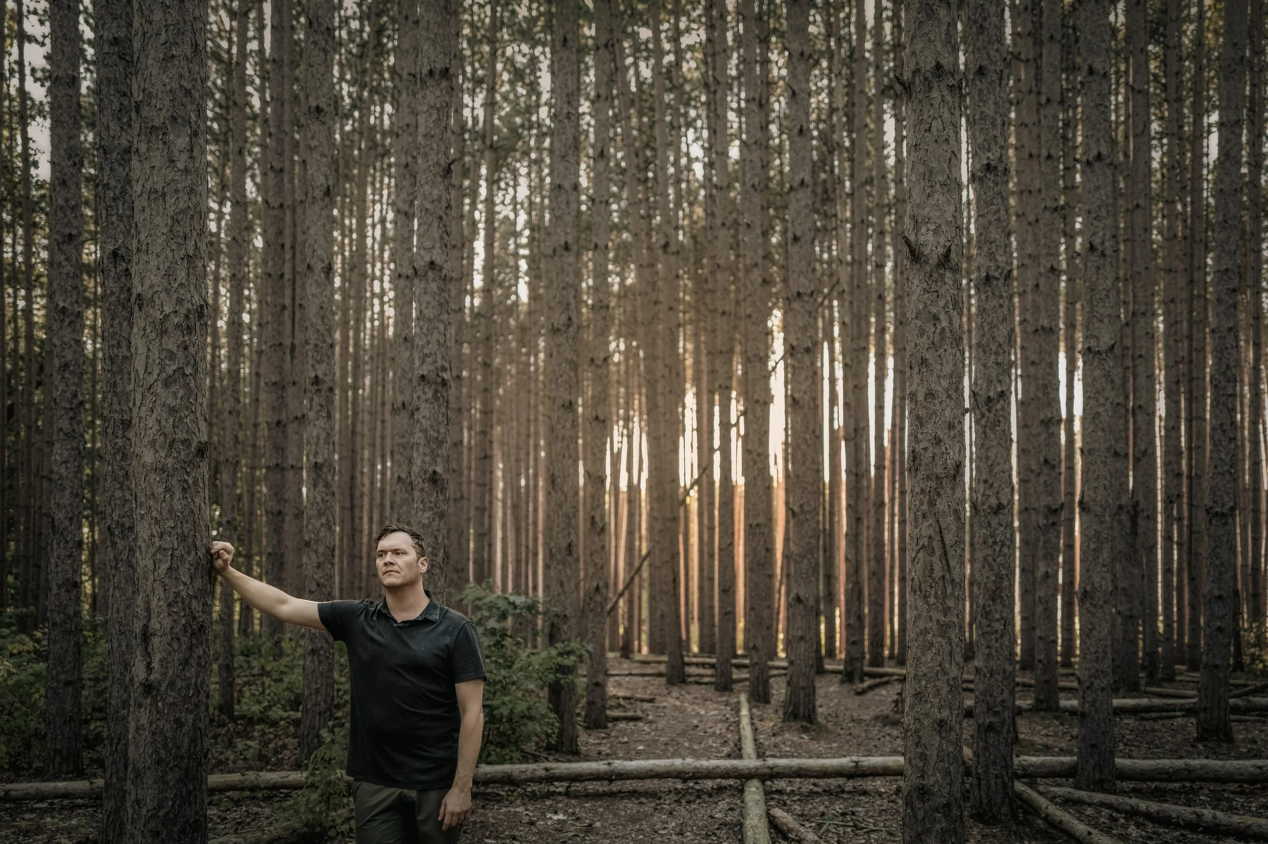 A man standing in a forest near a tree, with tall trees around him and sunlight filtering through the branches.