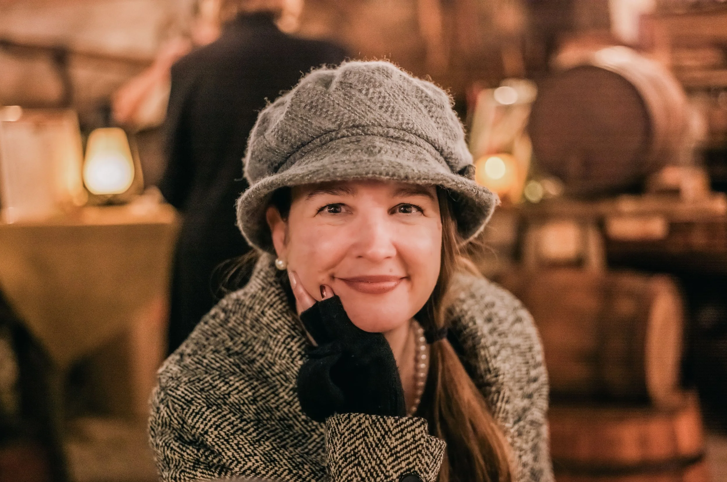 A woman smiling while sitting in a cozy, warmly lit indoor setting. She is wearing a gray hat, black gloves, a pearl necklace, and a patterned coat.