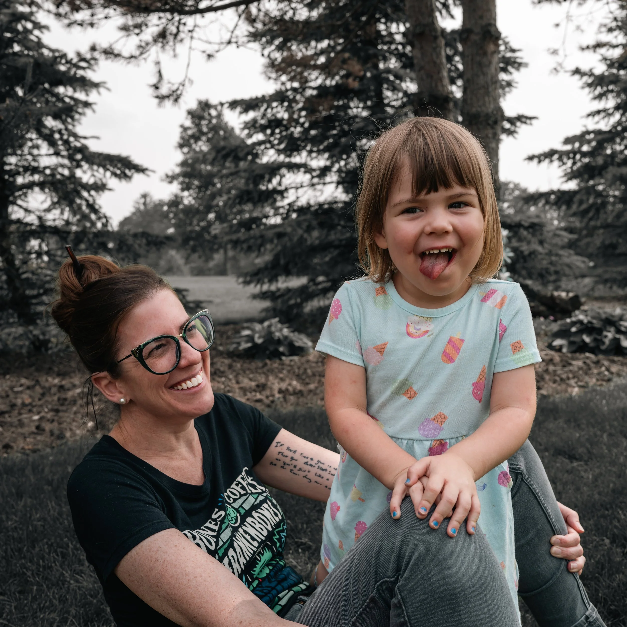 A woman with glasses and a black t-shirt smiling while sitting on the ground, holding a young girl on her lap, who is sticking out her tongue and making a playful face outdoors with trees in the background.