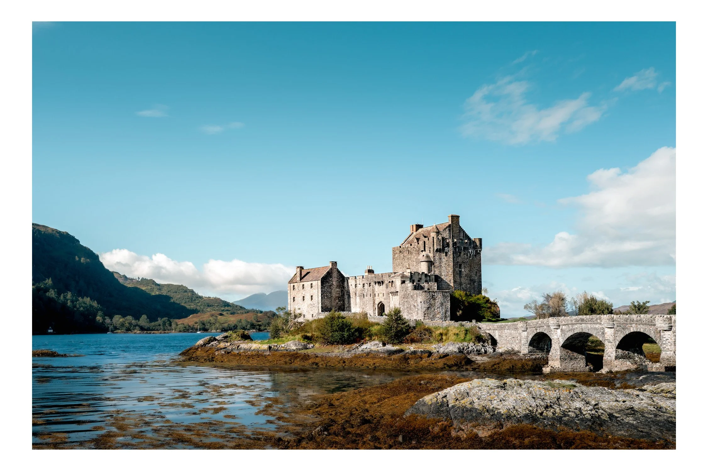 A historic stone castle located on a small island, connected to the mainland by a stone bridge, with water and mountains in the background under a blue sky with scattered clouds.
