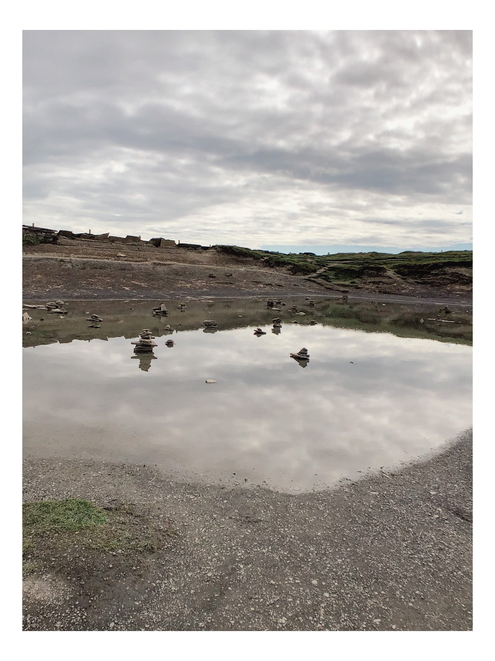A small natural pond with balance rocks stacked on top of each other in the water, reflecting the cloudy sky above.
