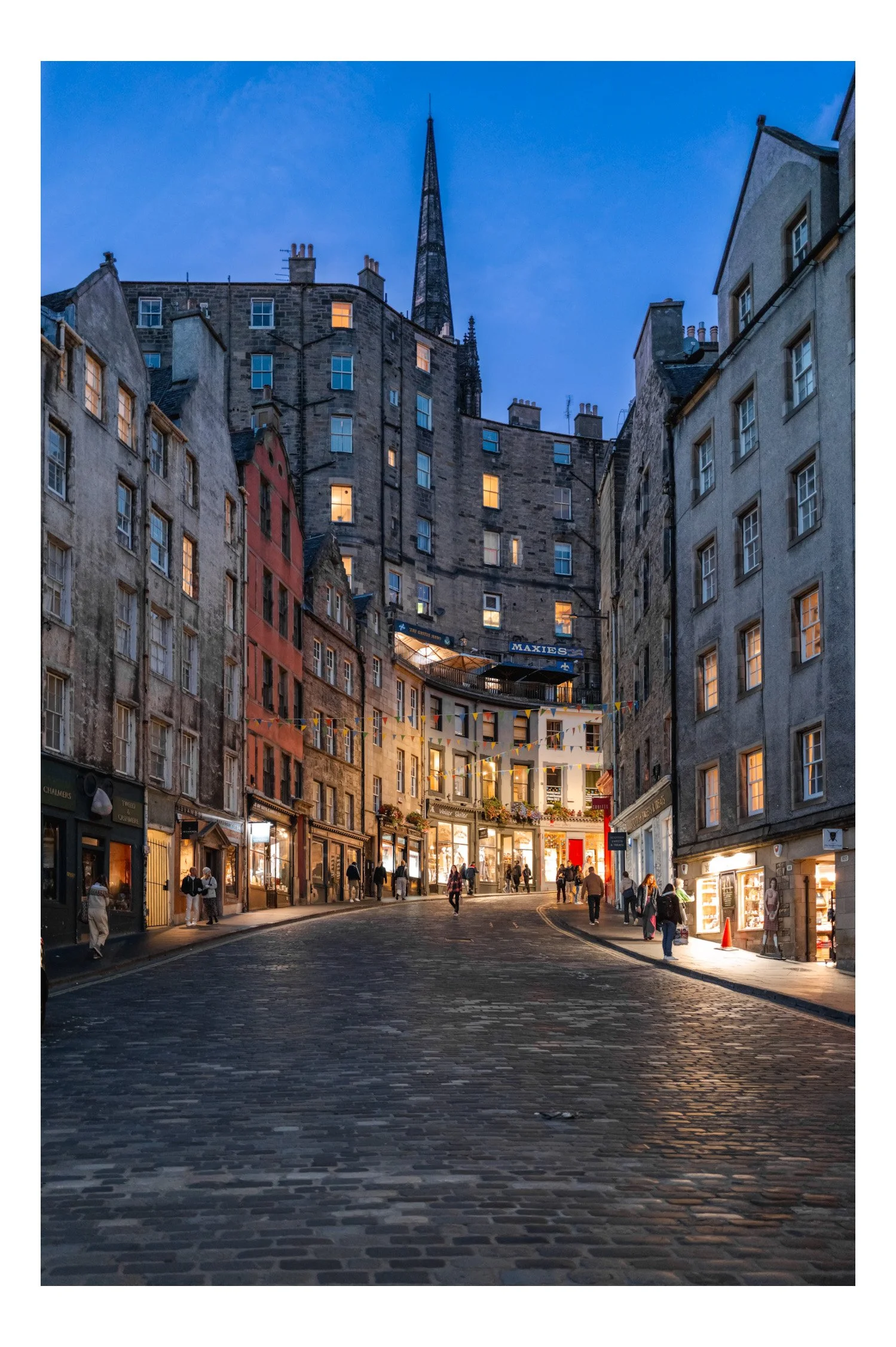 City street scene at dusk with cobblestone road, pedestrians, illuminated shopfronts, and historic buildings with a church steeple in the background.