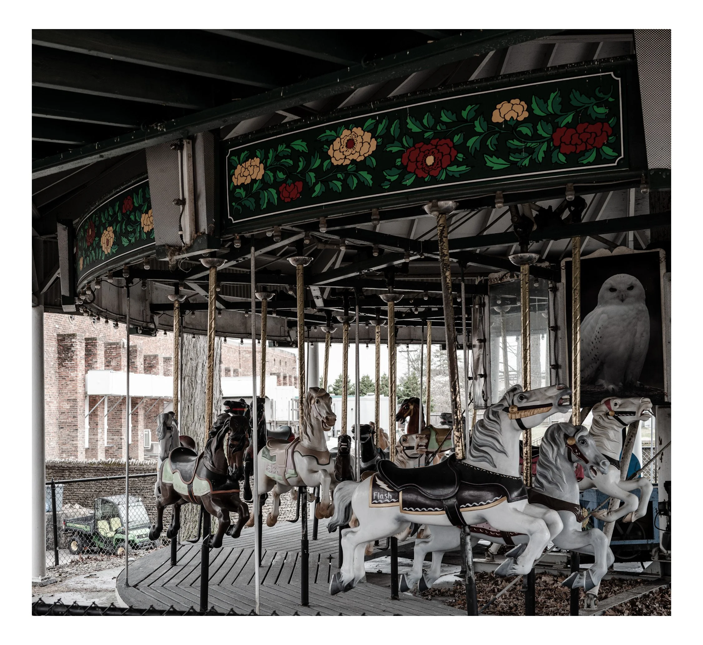 Empty vintage carousel with painted horses and owl picture, under a dark roof with floral decorations, outdoor setting with brick buildings and trees in background.