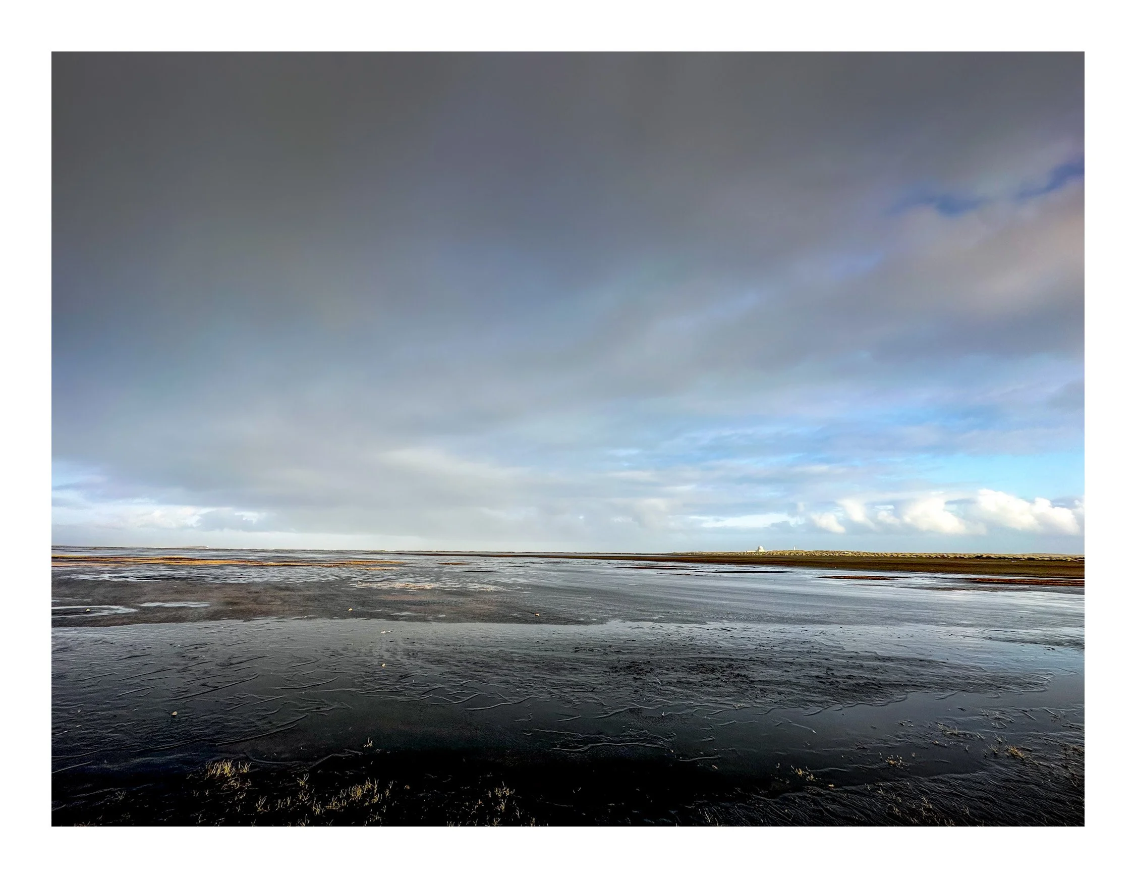 Overcast sky with patches of blue, view of a frozen, snow-covered landscape with cracks in the ice and a distant horizon with a small structure or building.