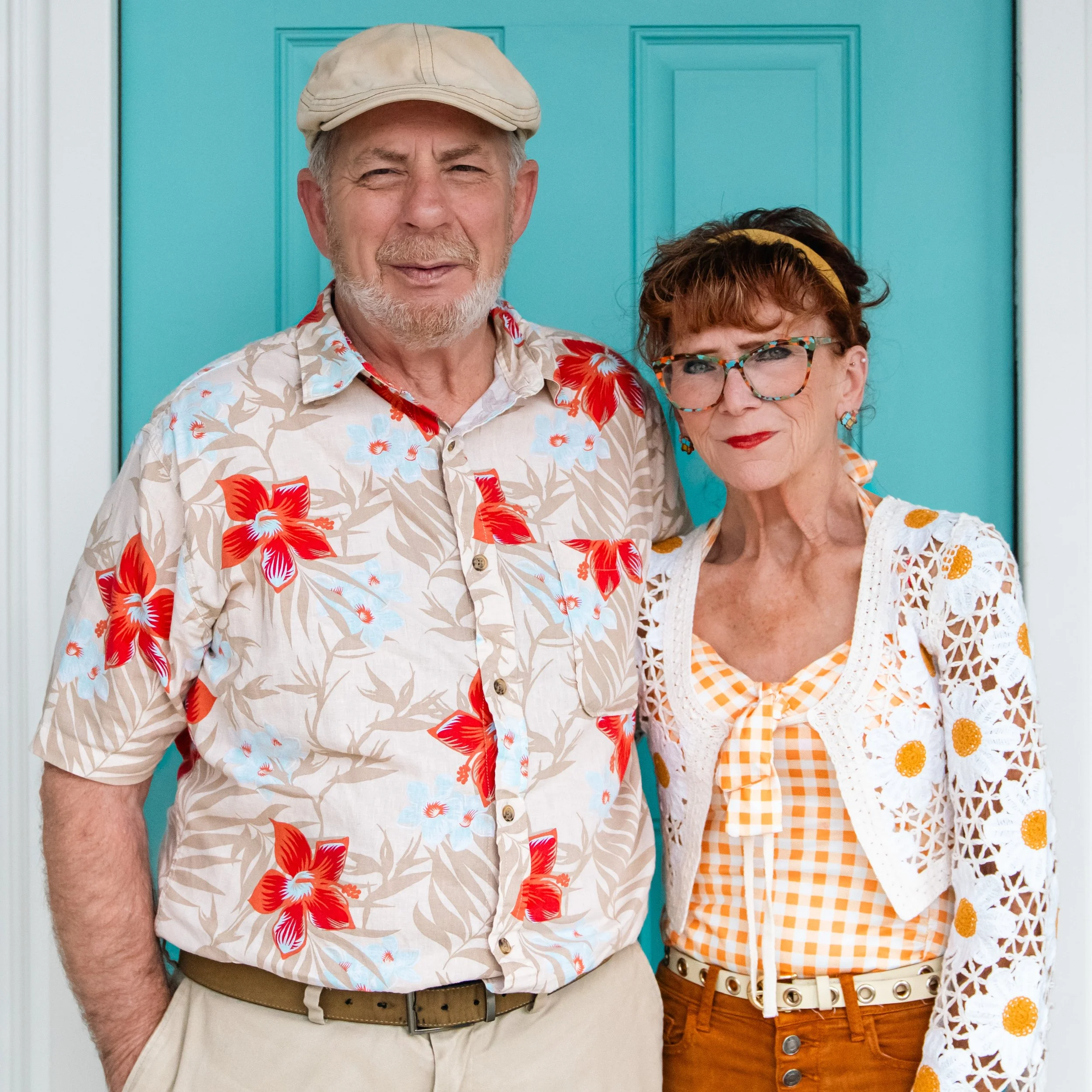 An elderly man and woman standing in front of a teal door, dressed in colorful, patterned summer clothes and smiling