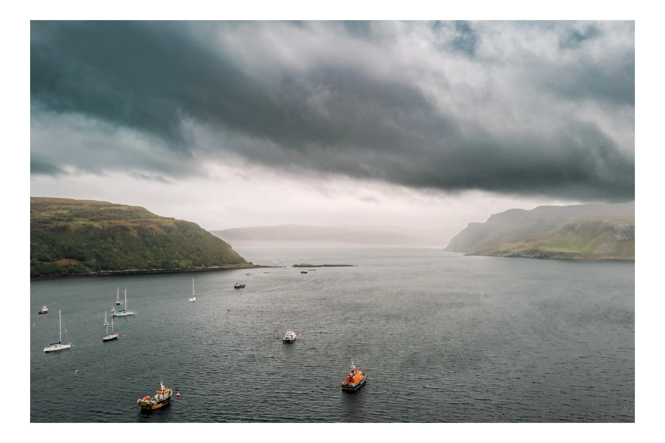 A misty harbor with several sailboats and a rescue boat floating on calm water, surrounded by green hills under a cloudy sky.