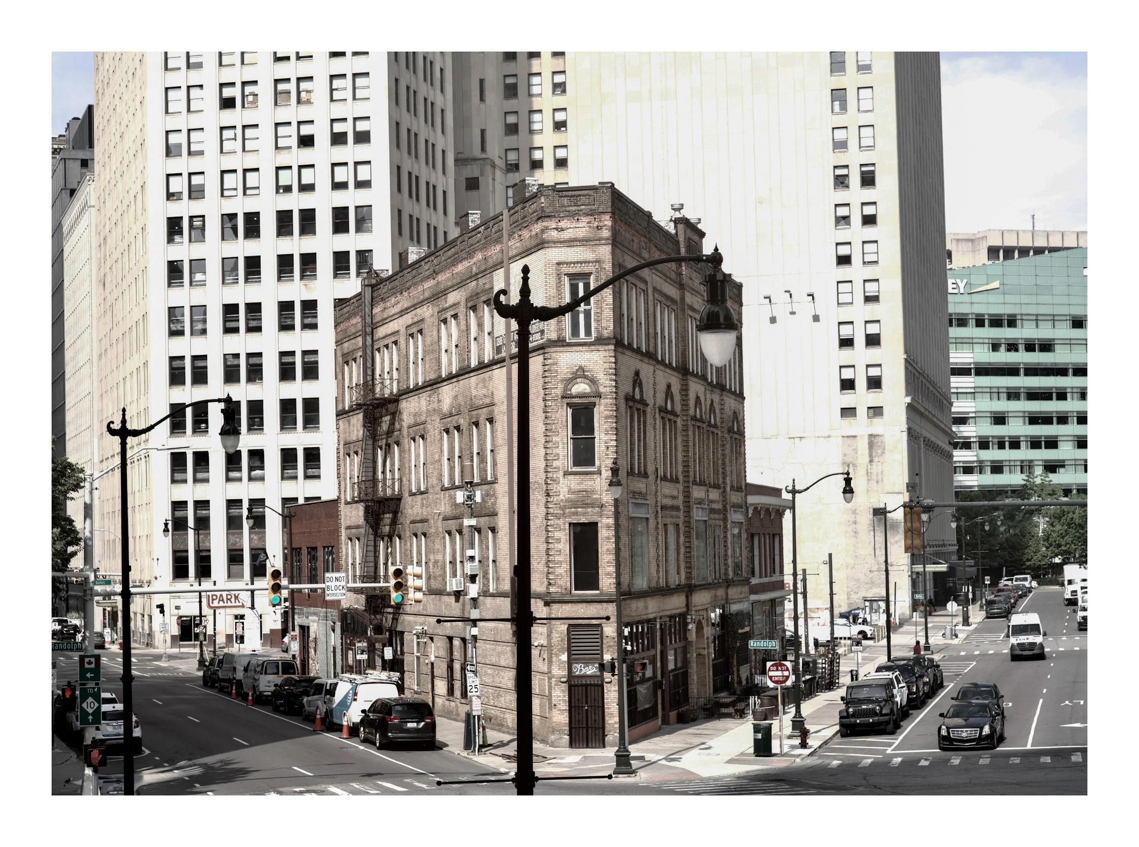 A city street corner with a historic brick building surrounded by modern high-rise buildings. parked cars, street lamps, and traffic lights are visible.