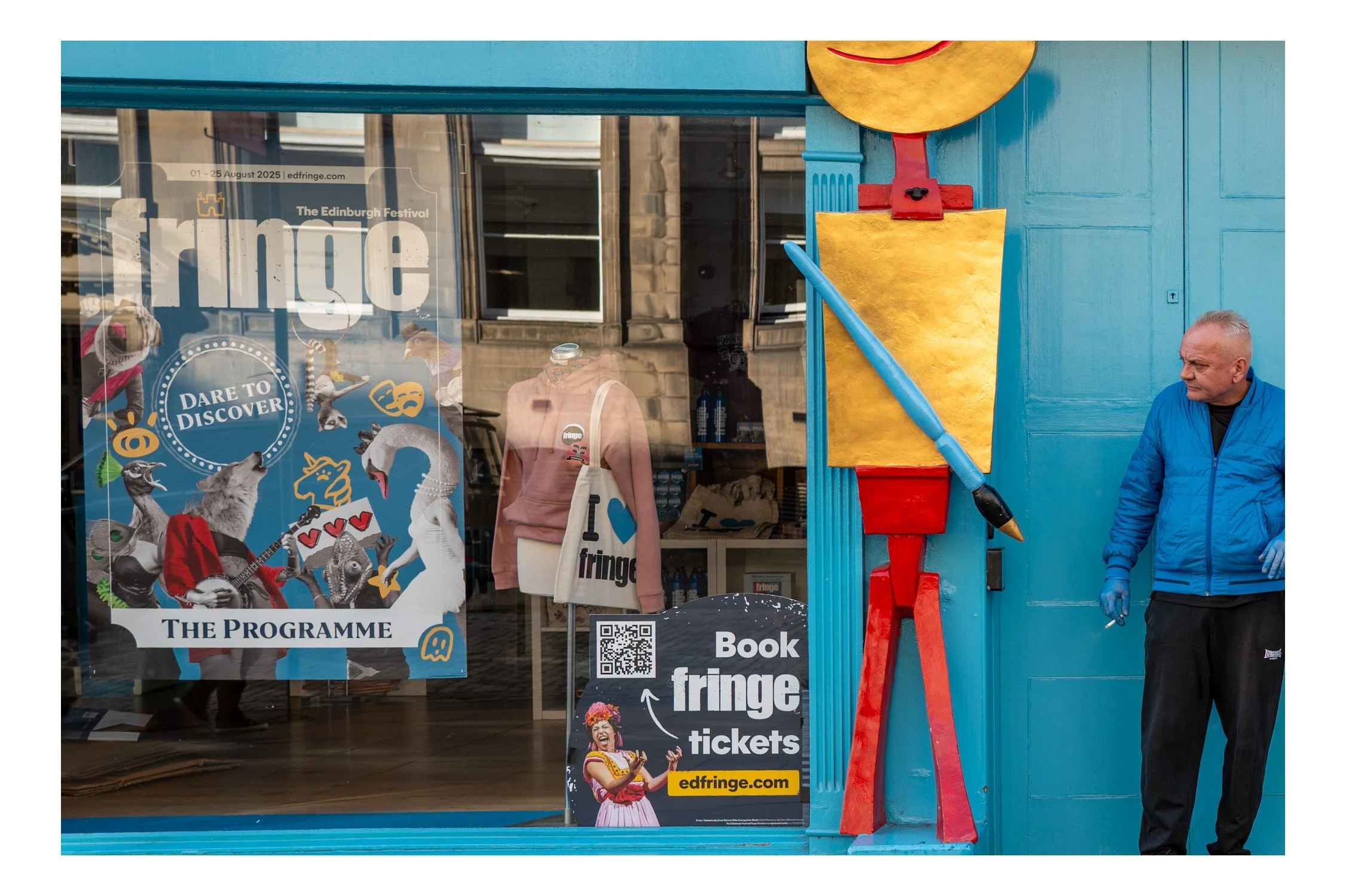 Street scene featuring a storefront decorated for the Edinburgh Festival with promotional posters and costumes. A man in a blue jacket stands next to a large, colorful sculpture of a paintbrush with a face.