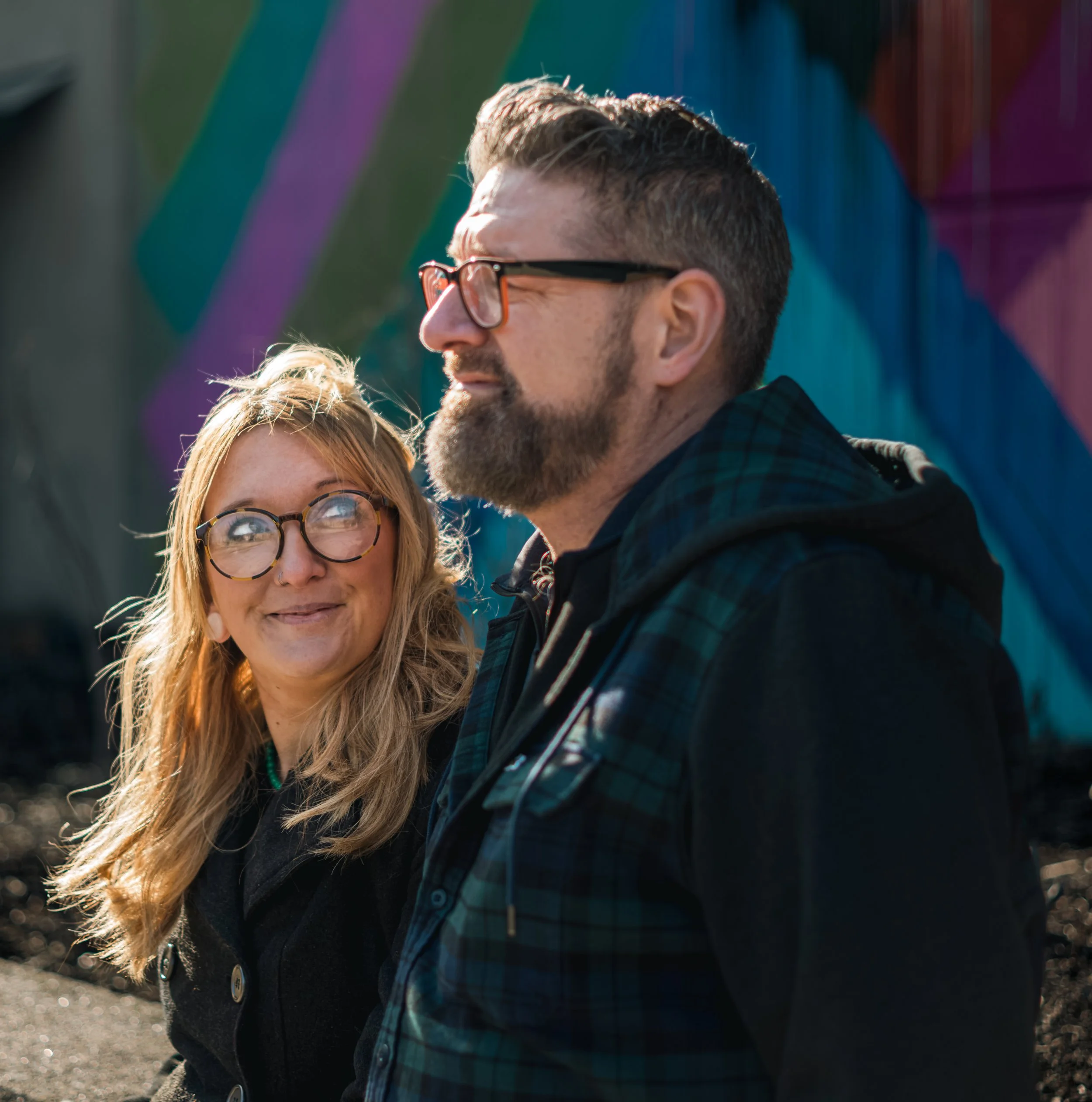 A woman with red hair and glasses looking at a man with glasses and a beard, both outdoors in front of a colorful mural.