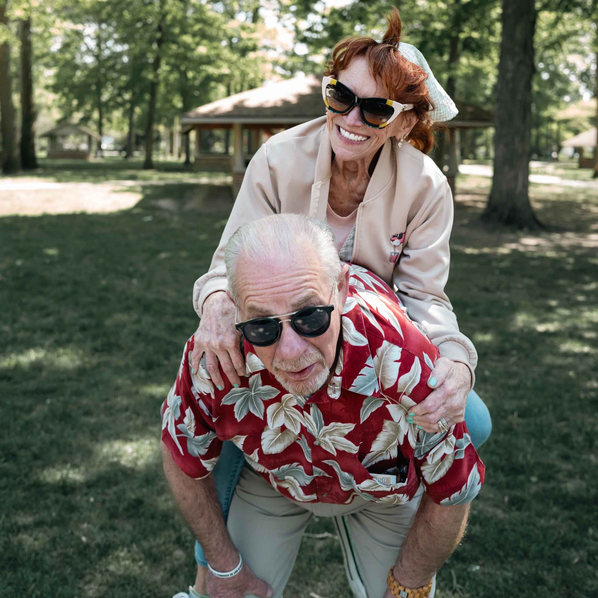 An elderly man wearing sunglasses and a red floral shirt is crouching outdoors, with an elderly woman with red hair, large sunglasses, and a beige jacket on his back. They are in a park with green grass and trees.