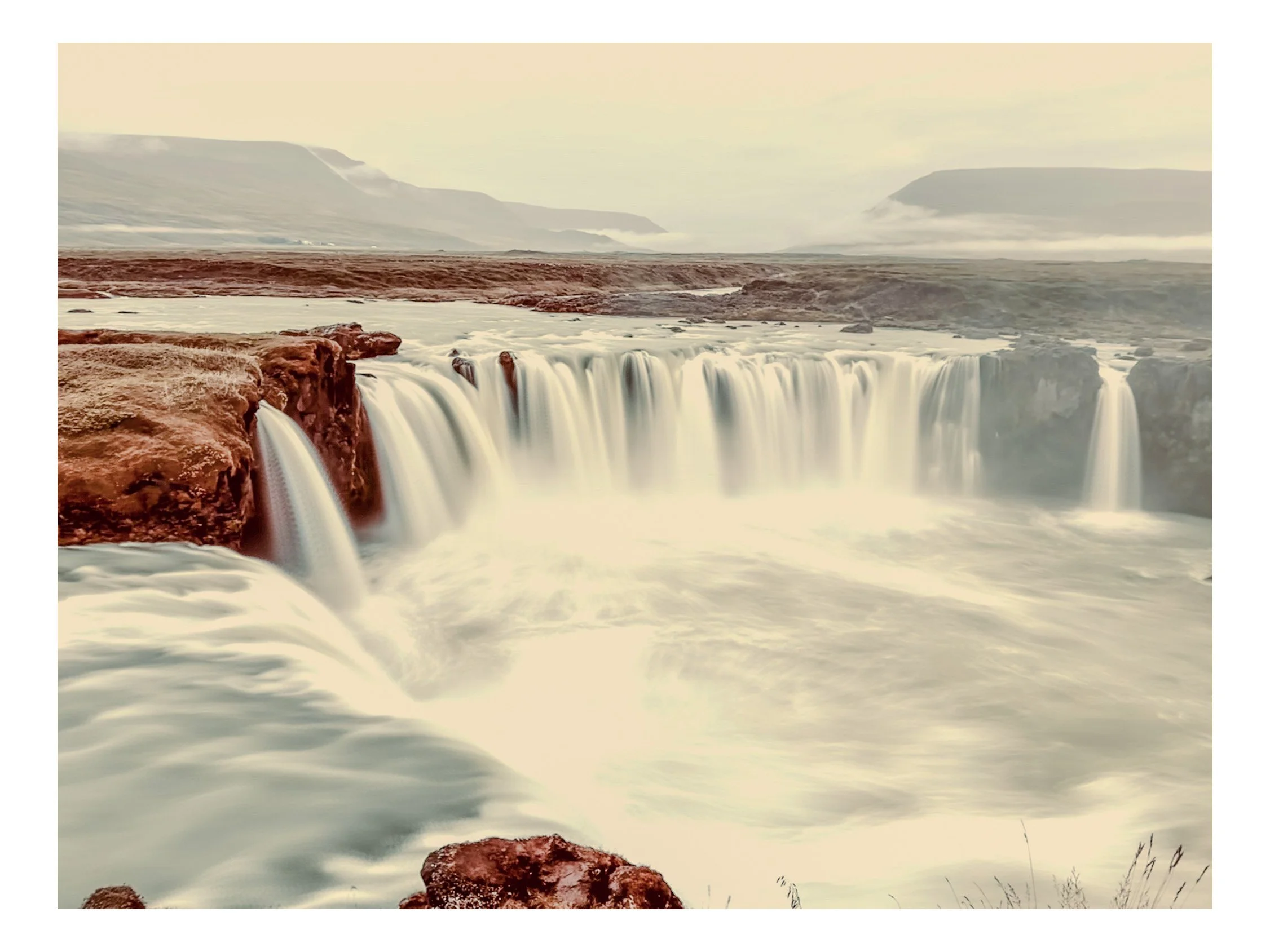 Waterfalls cascading over reddish-brown rocks in a wide river, with mist and distant mountains under a cloudy sky.