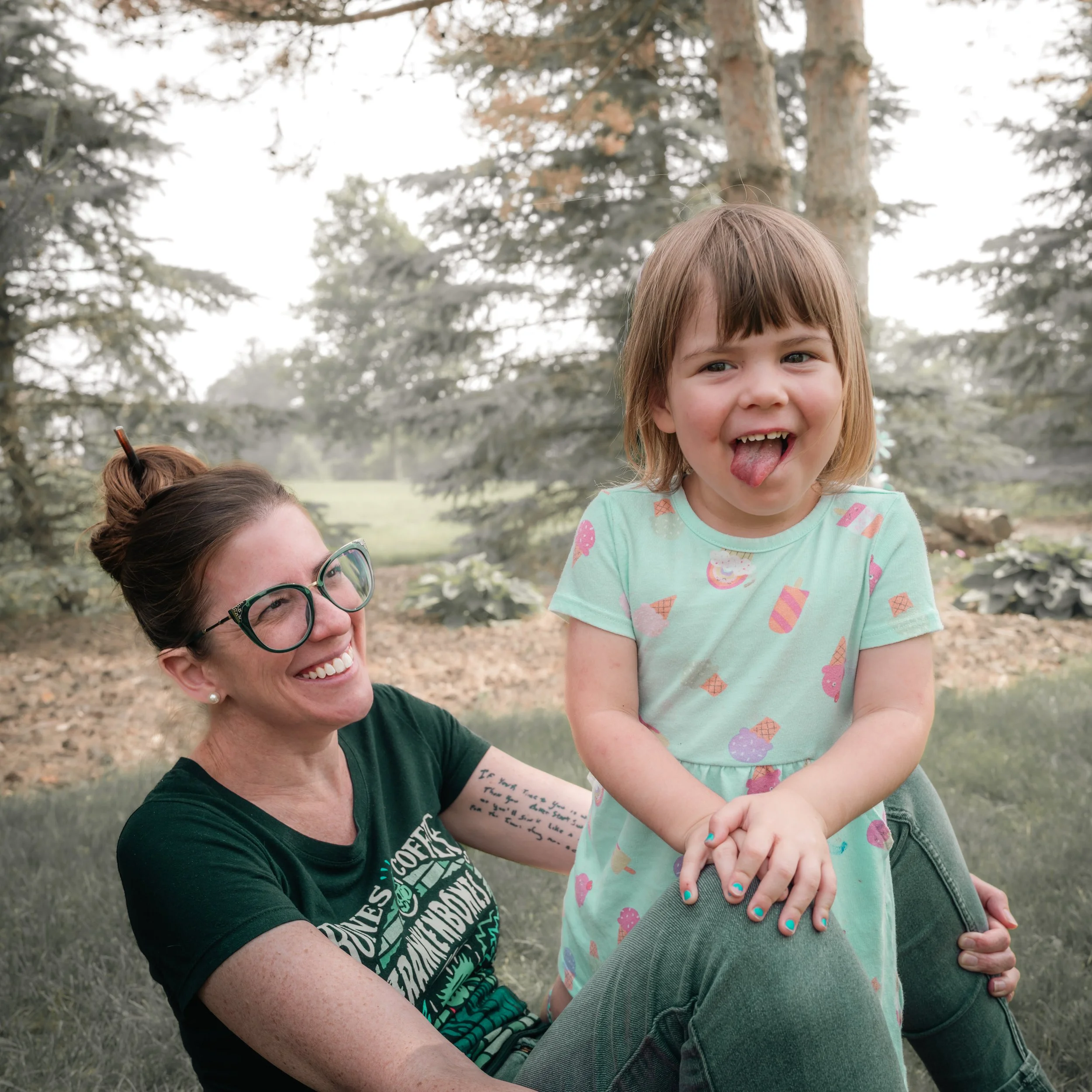 A woman with glasses and a woman tattoo on her arm, smiling at a young girl with short light brown hair sticking out her tongue, sitting on her lap outdoors surrounded by trees and greenery