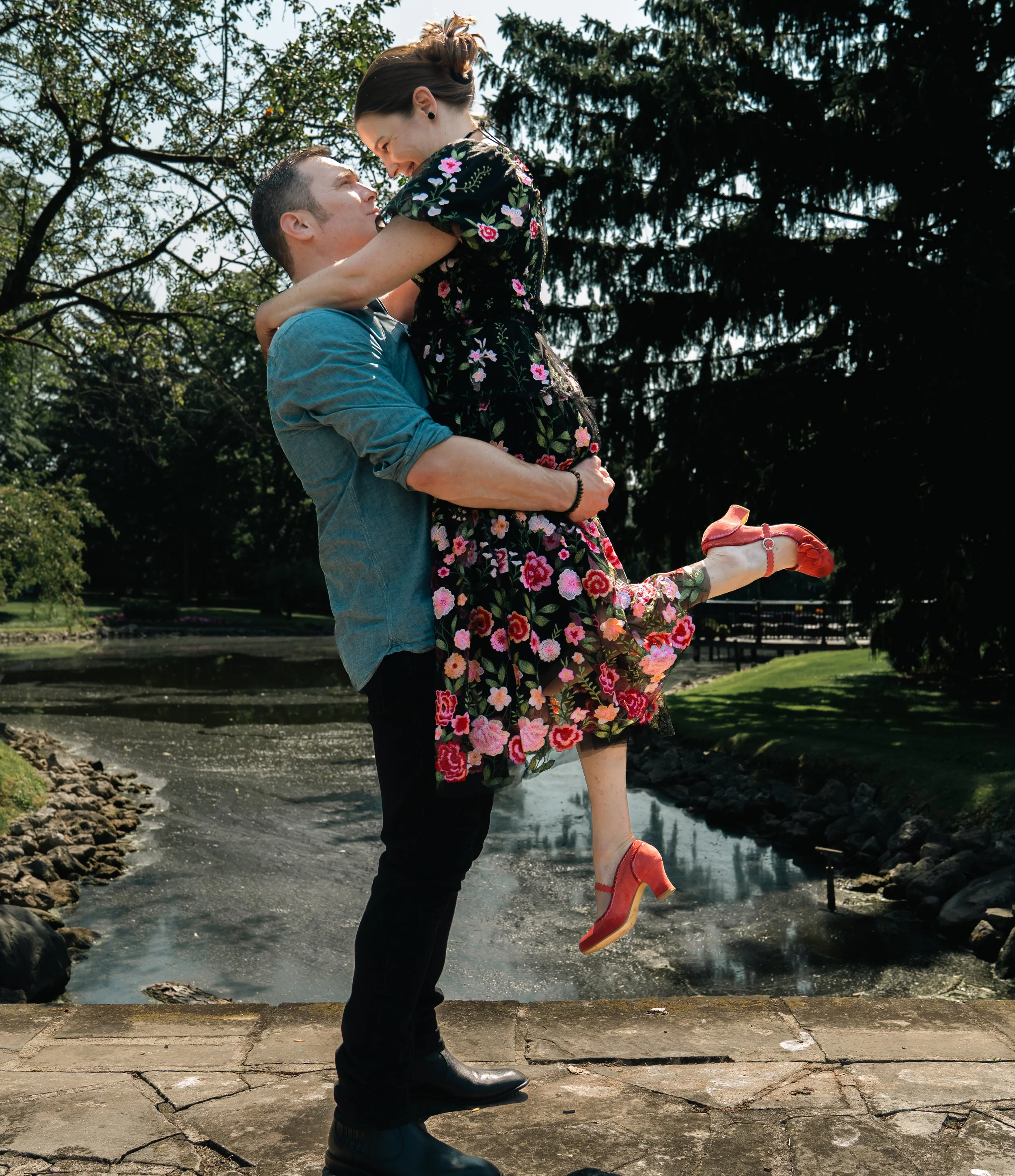 A man lifting a woman in a floral dress and red shoes near a pond in a park.