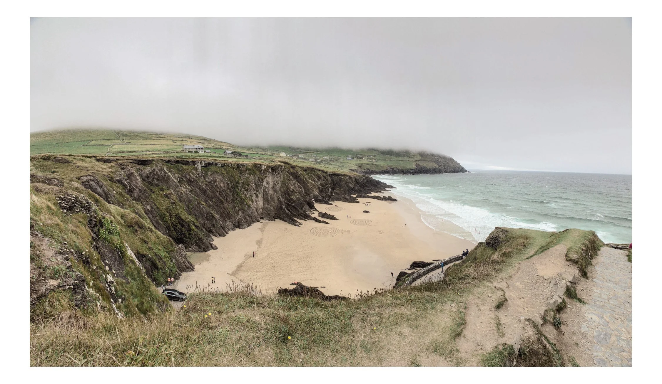 A panoramic view of a beach with cliffs, sandy shore, and waves, under a cloudy, foggy sky.
