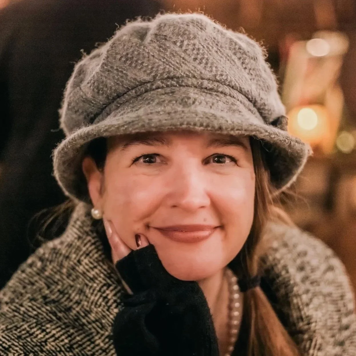 A woman wearing a gray wool hat, pearl earrings, and a tweed coat, smiling softly with her chin resting on her hand, in a warmly lit indoor setting.