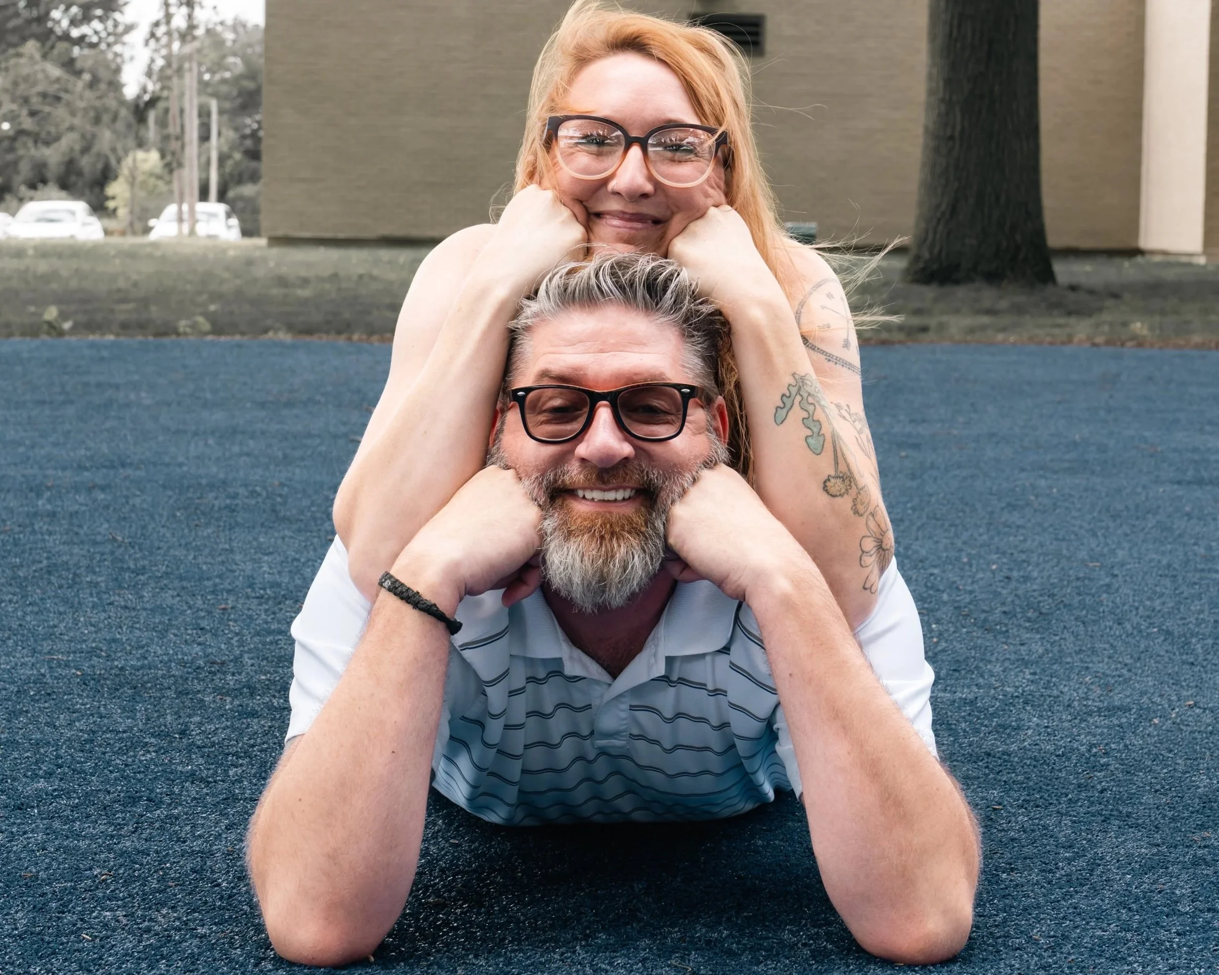 A man and woman with glasses relaxing outdoors. The man is lying on his stomach on a blue surface, smiling with his hands supporting his face. The woman is leaning on his back, resting her chin on her hands, smiling. They are in a park with trees and a building in the background.