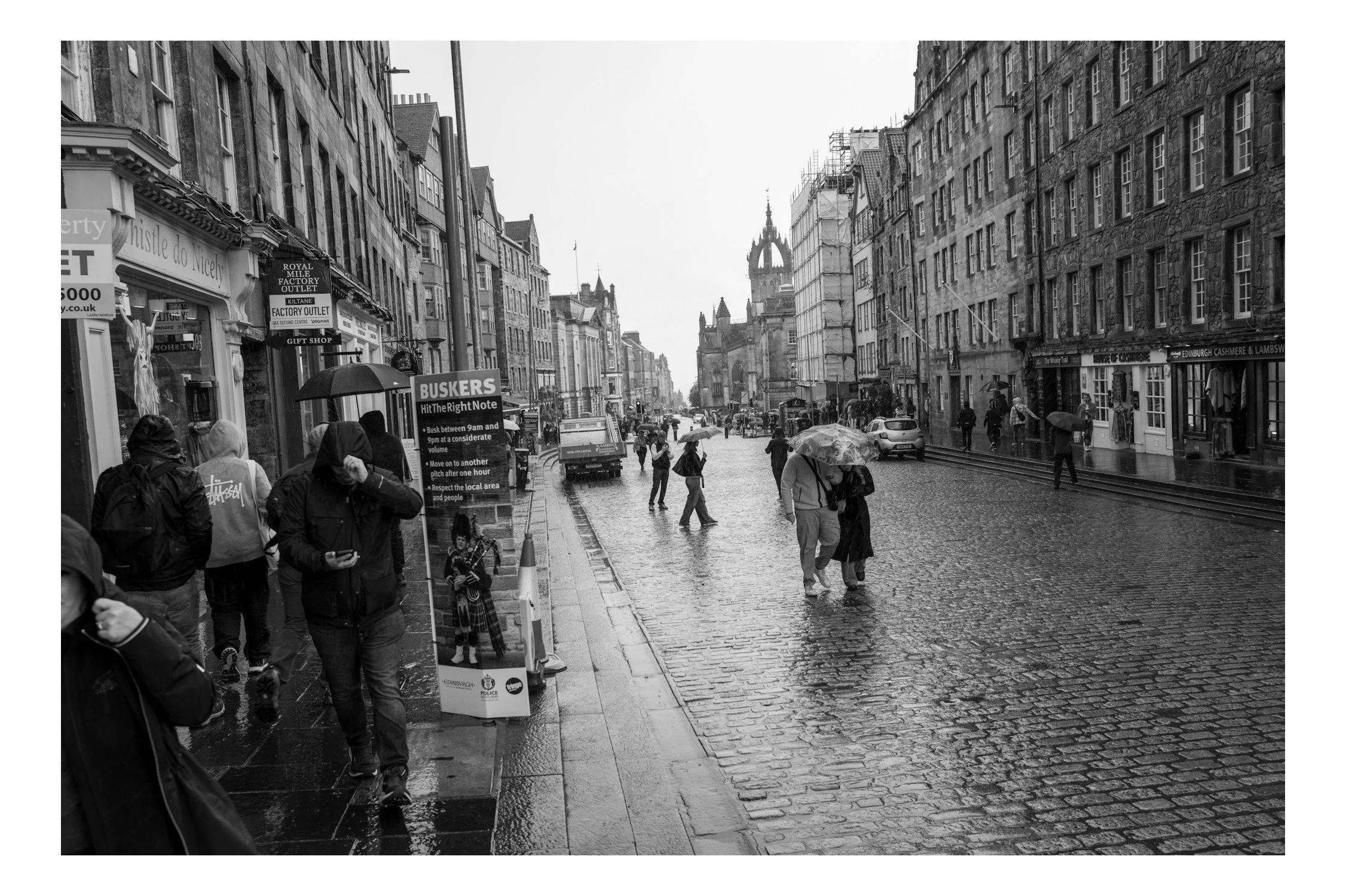 People walking on a wet cobblestone street with umbrellas on a rainy day in an old city. Buildings with shops and signs line the street, and a historic church or cathedral is visible in the background.