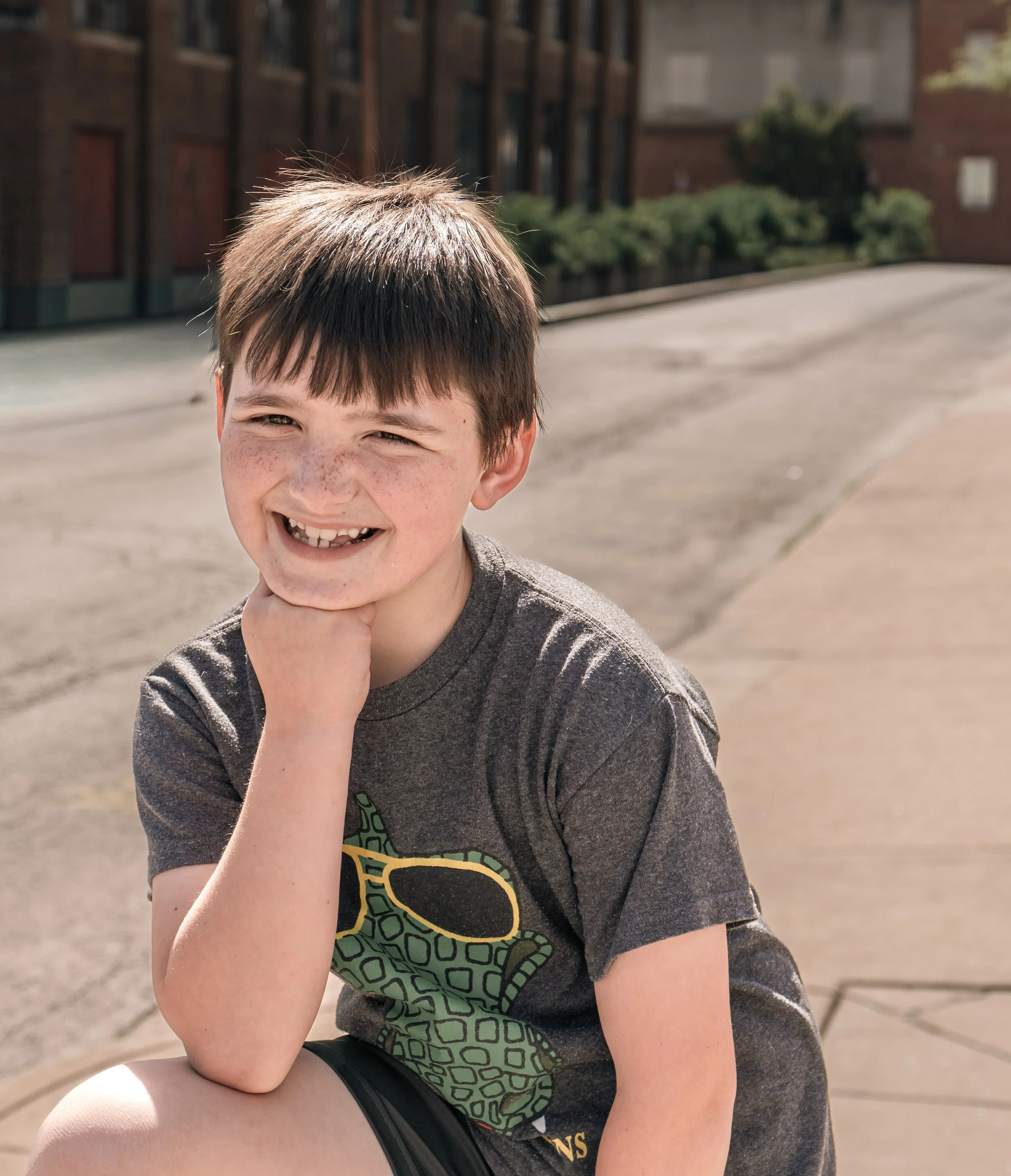 A boy with brown hair, freckles, and a big smile, resting his chin on his hand outdoors in bright sunlight.