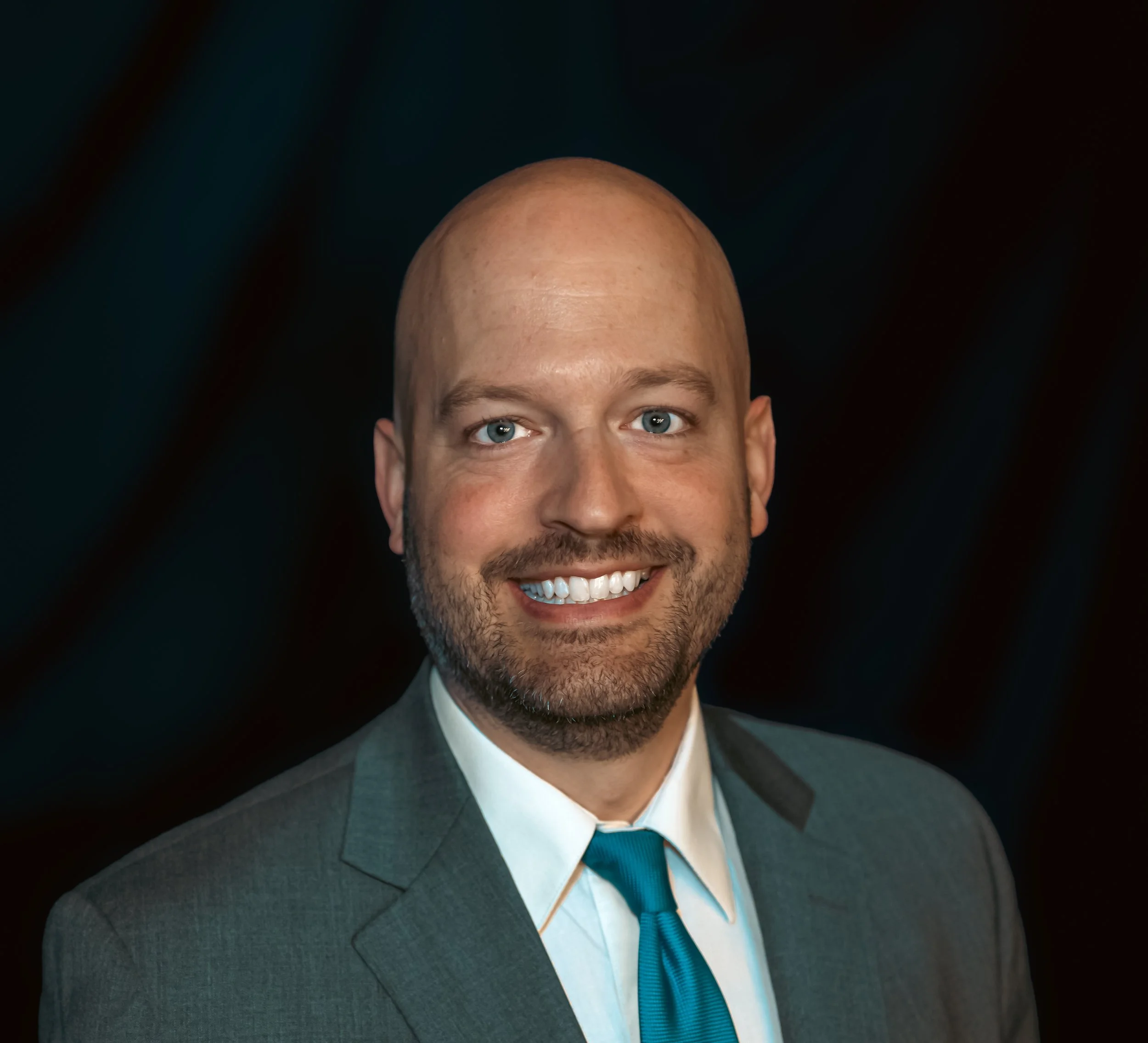 Headshot of a smiling man with blue eyes, a bald head, and facial hair, wearing a gray suit, white shirt, and blue tie against a dark background.