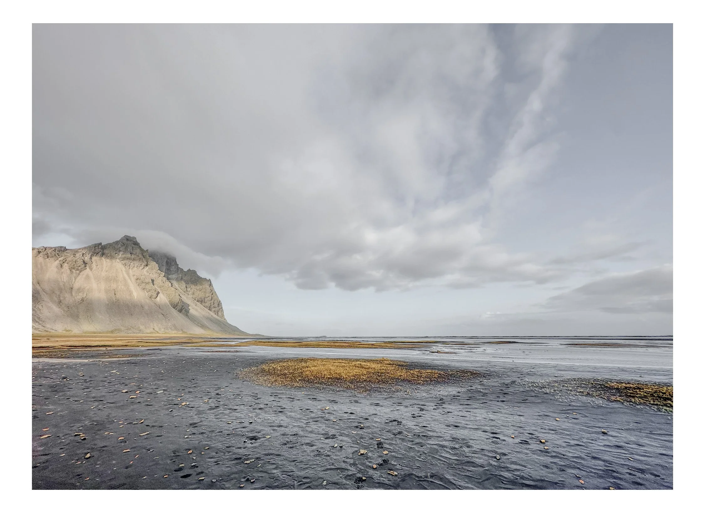 A vast coastal landscape featuring a black sand beach, distant mountains with steep cliffs, and partly cloudy skies overhead.