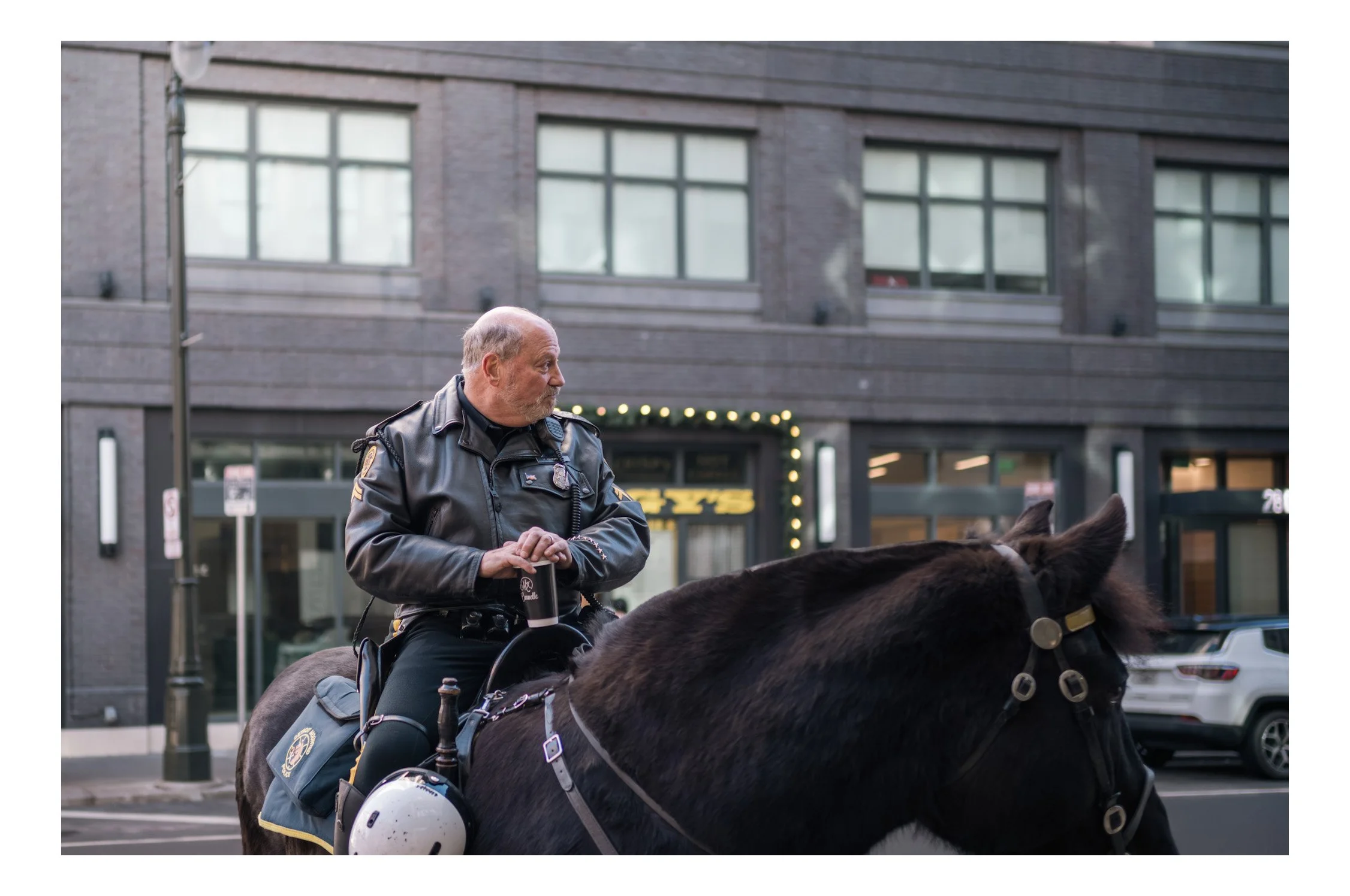 A police officer riding a horse on a city street, holding a coffee cup, with buildings and cars in the background.