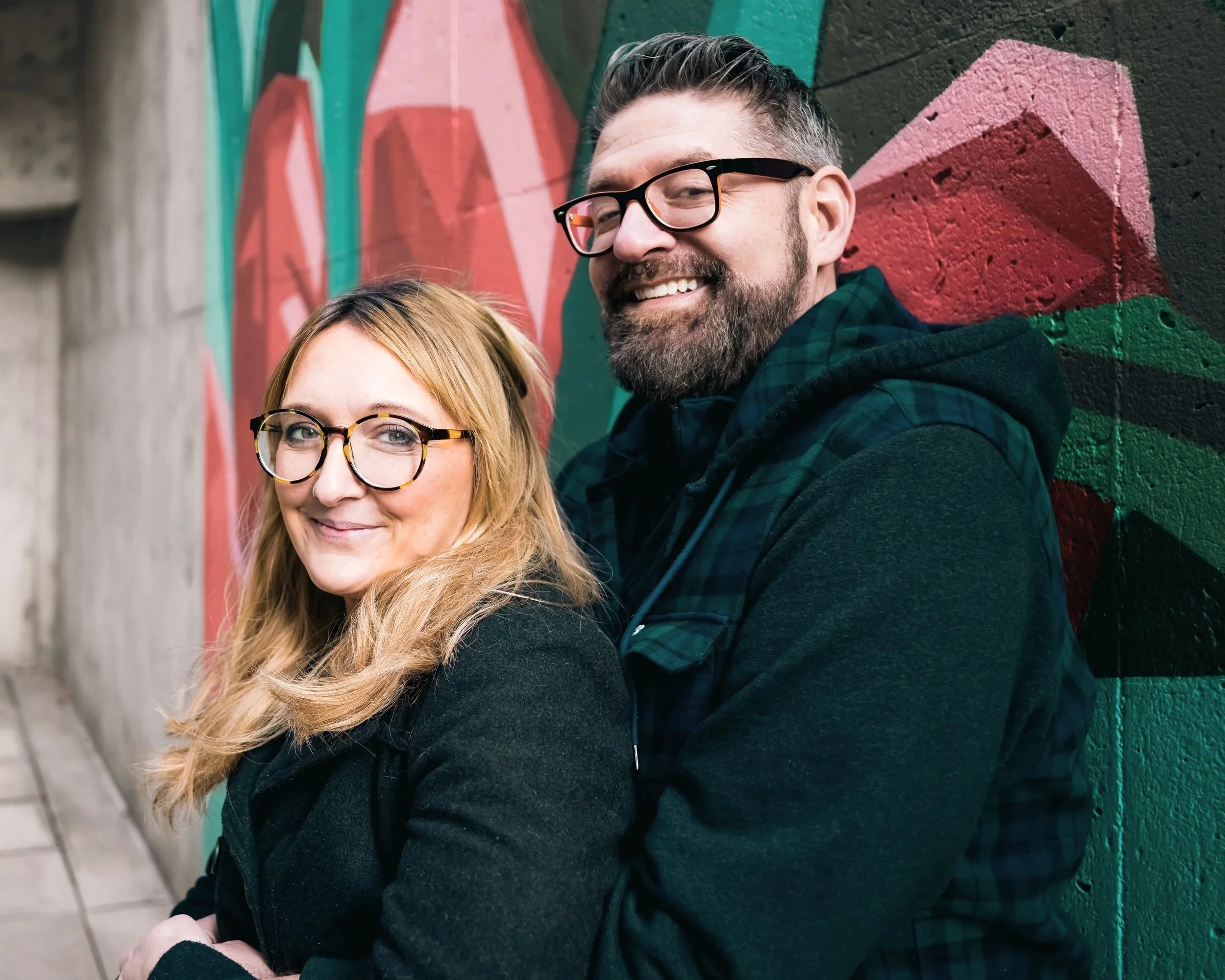 A smiling woman and man wearing glasses standing against a colorful graffiti wall.