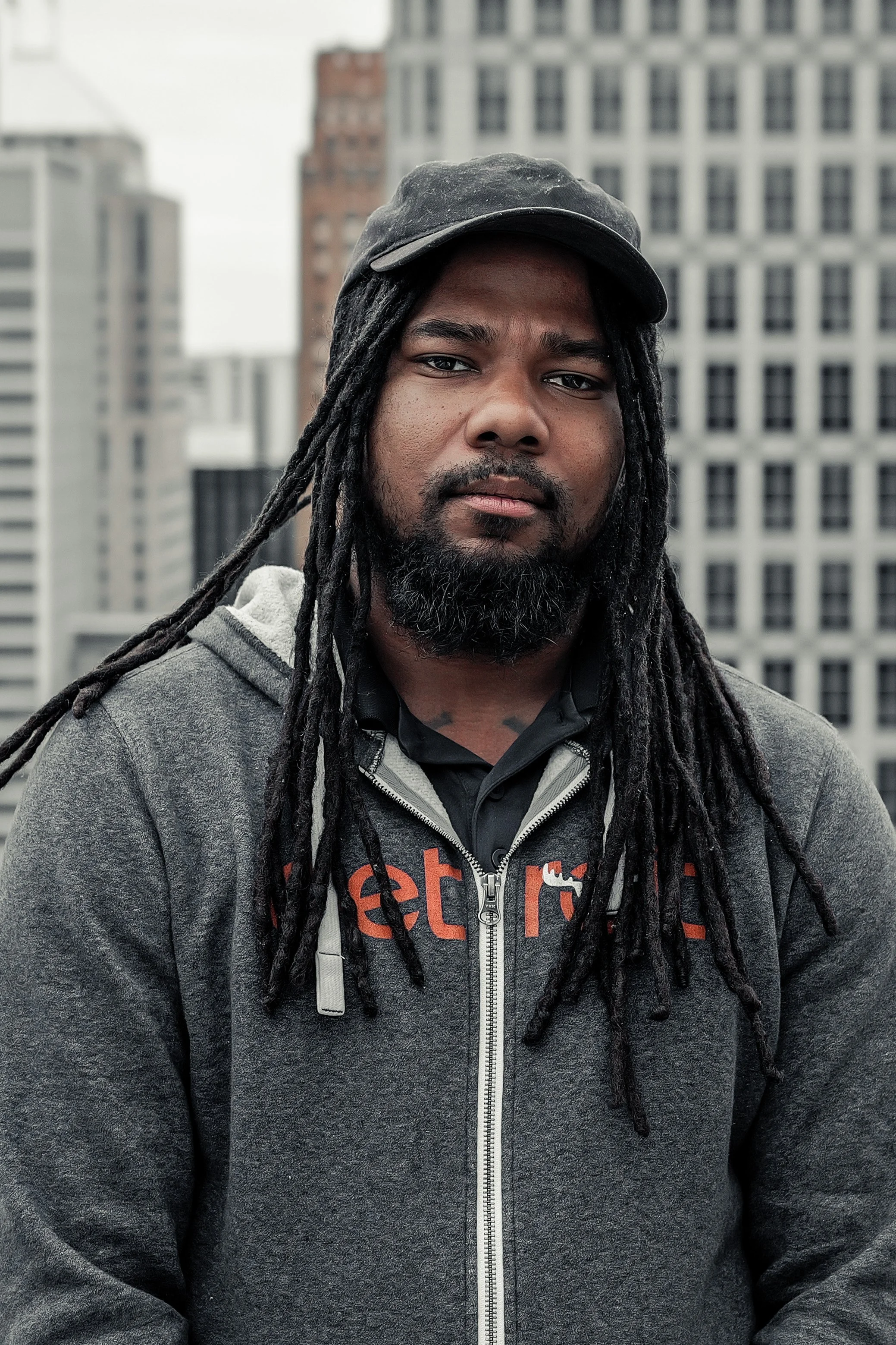 A man with long dreadlocks and a beard wearing a dark baseball cap and a gray zip-up hoodie, standing outdoors in front of tall city buildings.